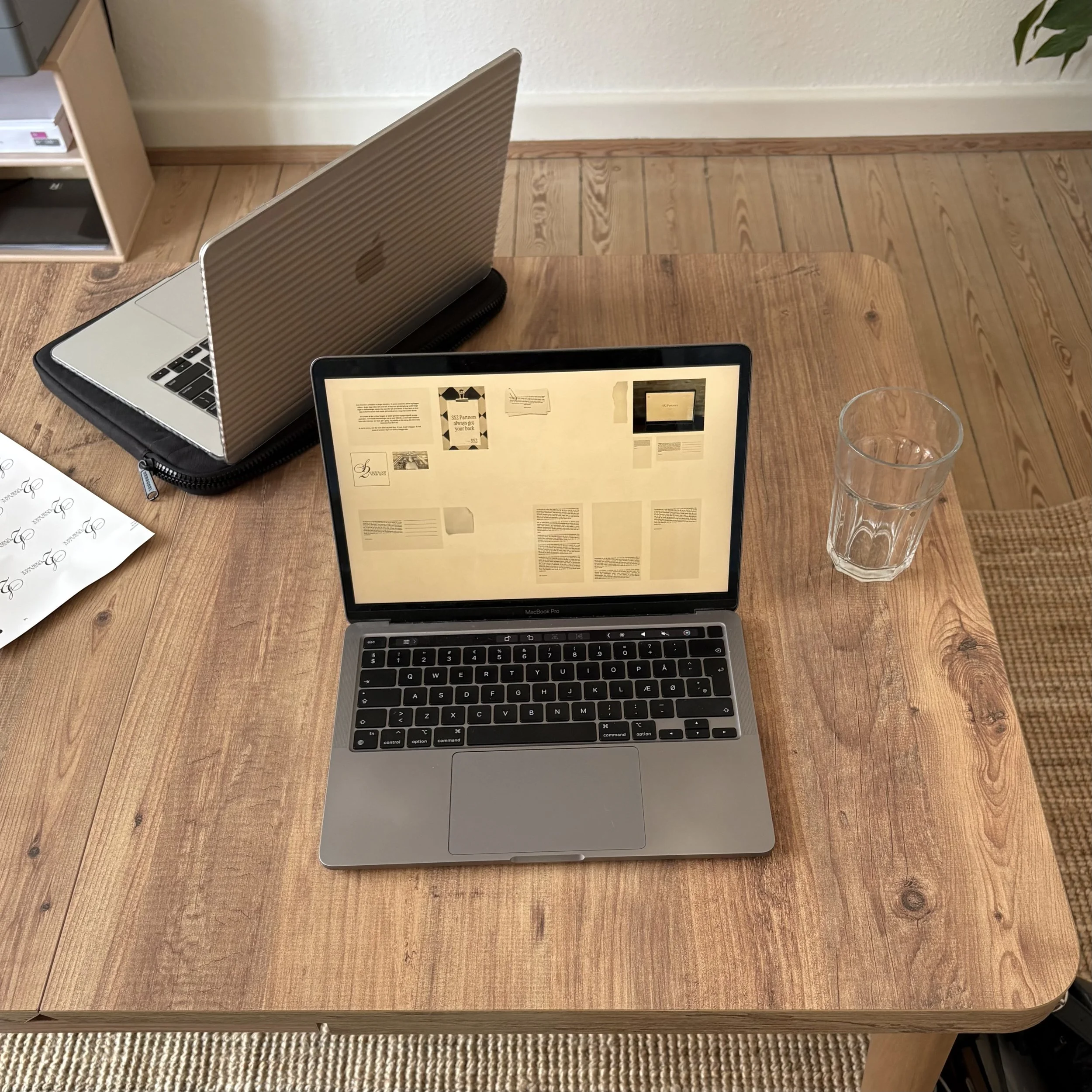 Et træbord med en åben MacBook Pro, en iPad, tre tomme vandglas, en notebook med papirark, en sort taske og et lille hyldesystem med bøger i baggrunden.