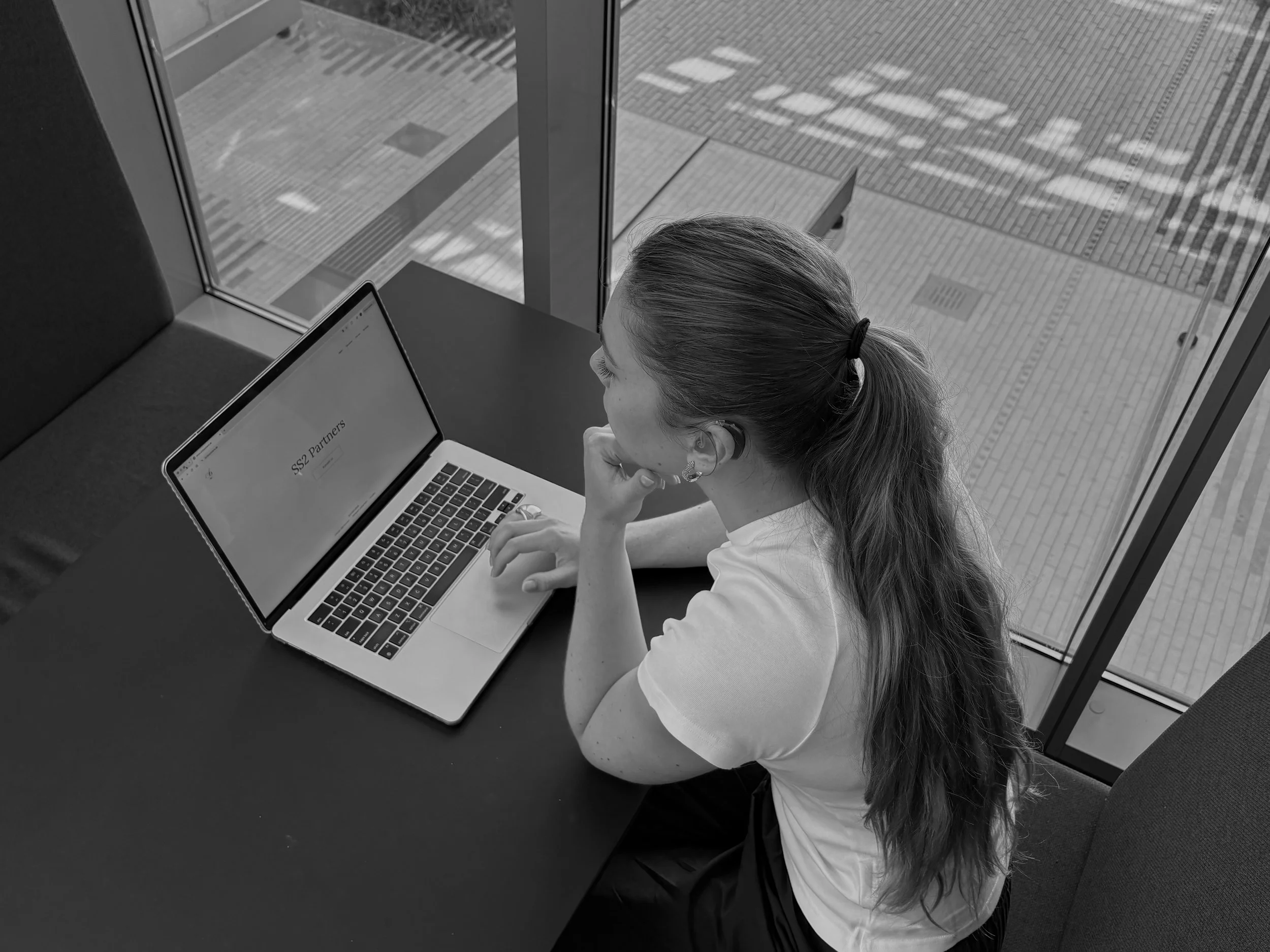 En kvinde med langt hår i en hestehale sidder ved et bord med en bærbar computer, der viser teksten 'SS2 Partners', og ser ud ad vinduet ind til en gade.