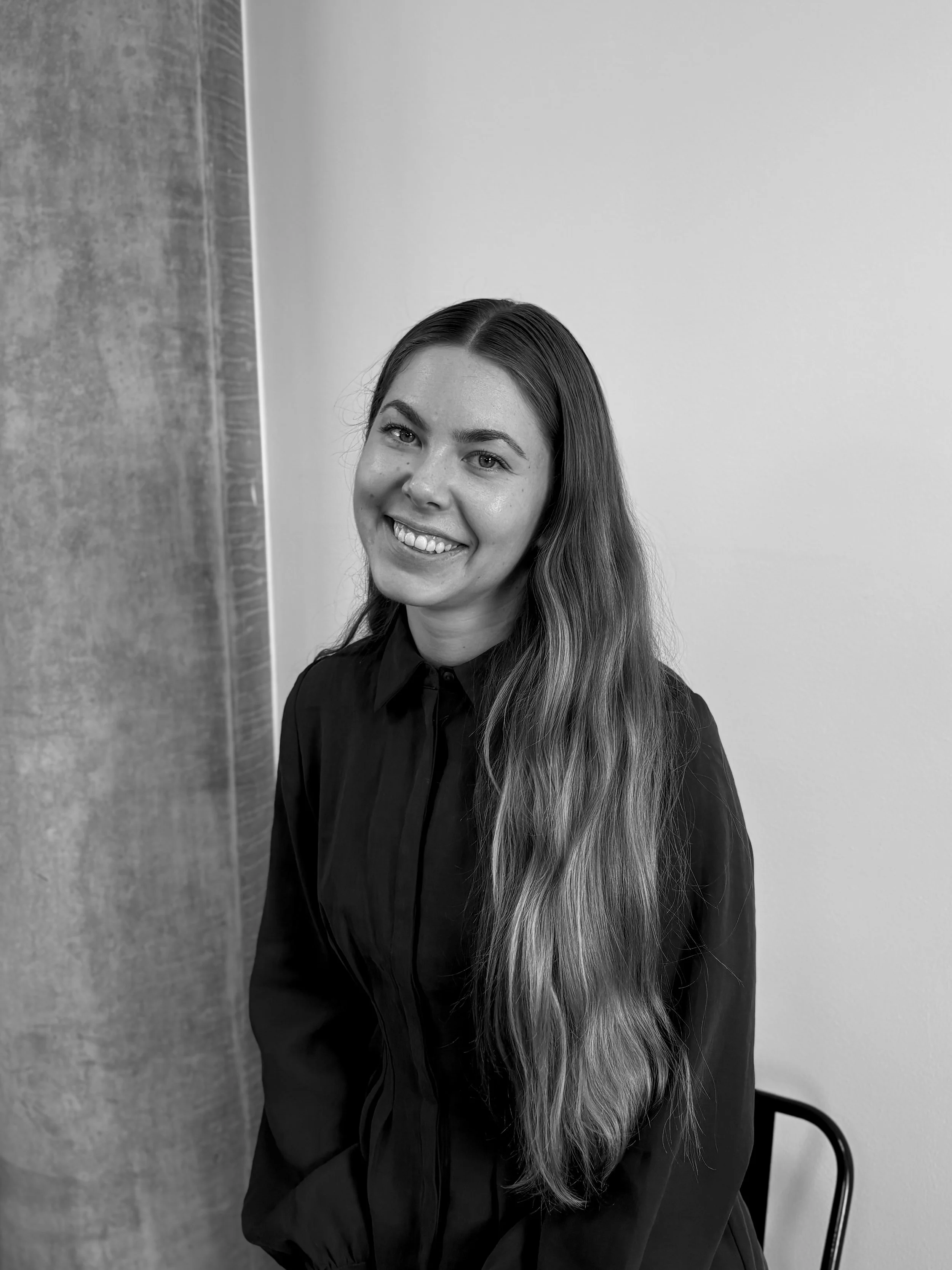Sort-hvid portrætfoto af en smilende ung kvinde med langt, bølget hår, iført en sort skjorte, siddende på en sort stol mod en enkel baggrund