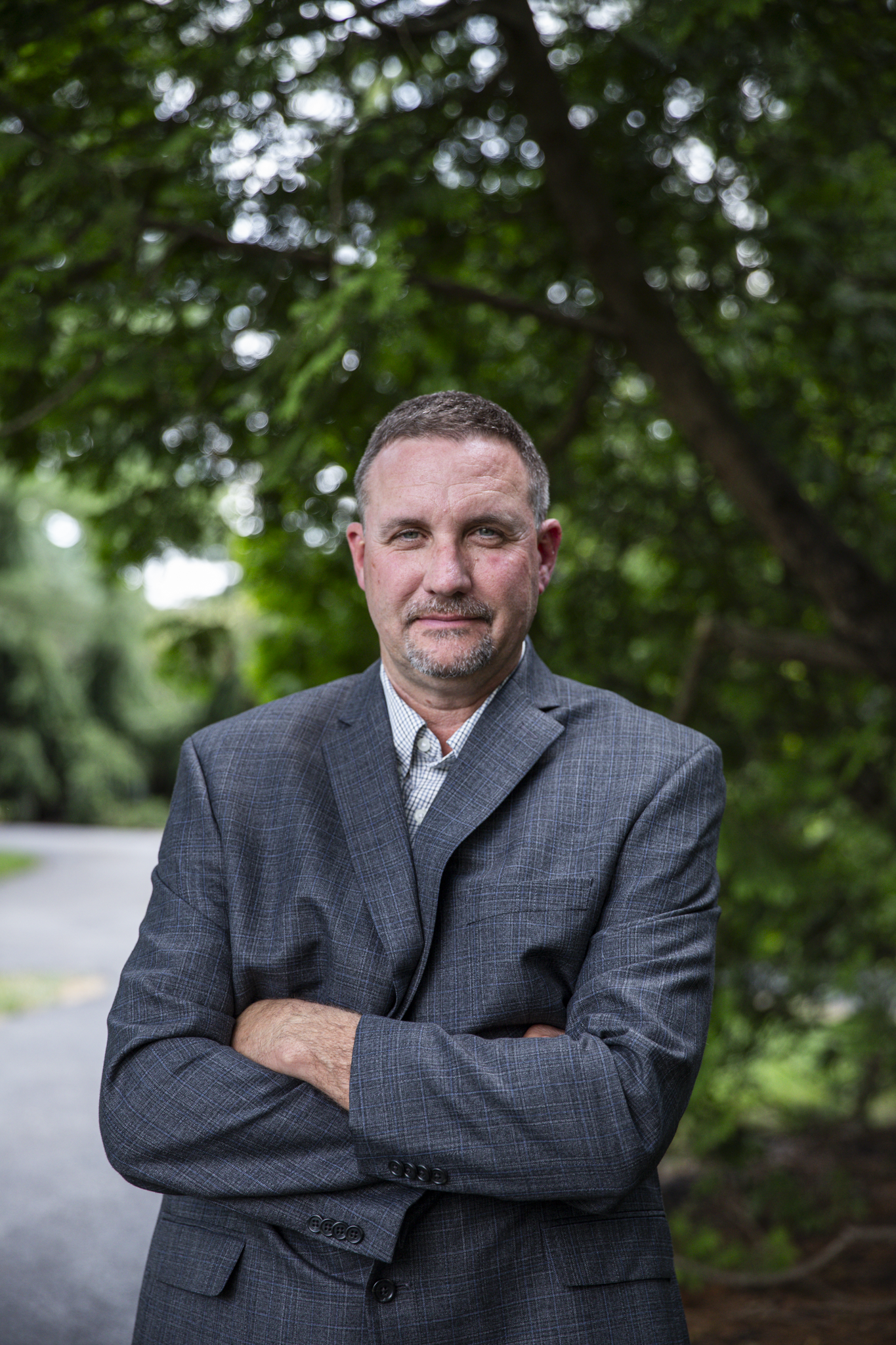A middle-aged man in a checkered suit jacket stands outdoors with his arms crossed in front of a lush green background of trees.