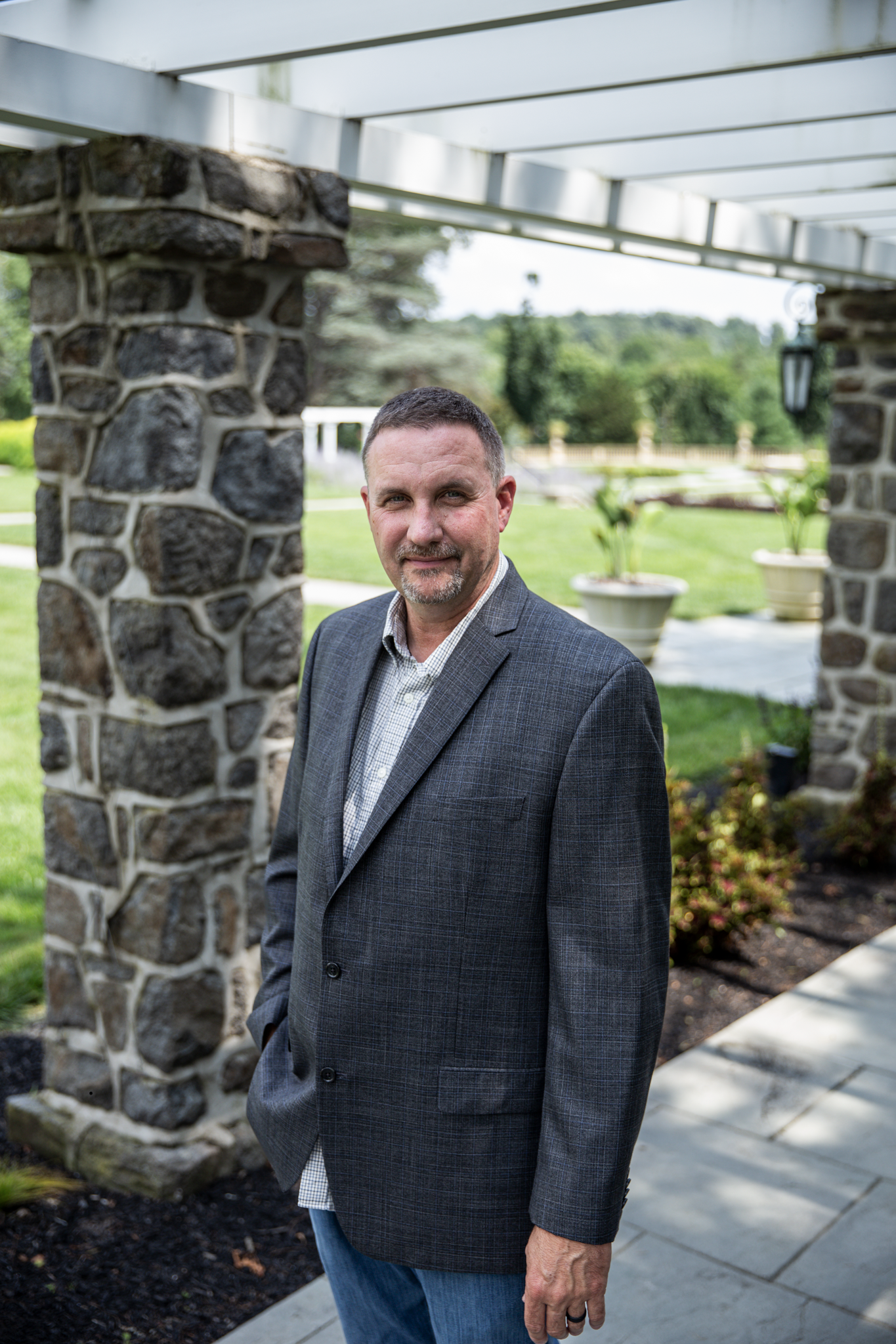 A middle-aged man with short dark hair and a beard, dressed in a checked blazer, standing outdoors near stone columns with greenery in the background.