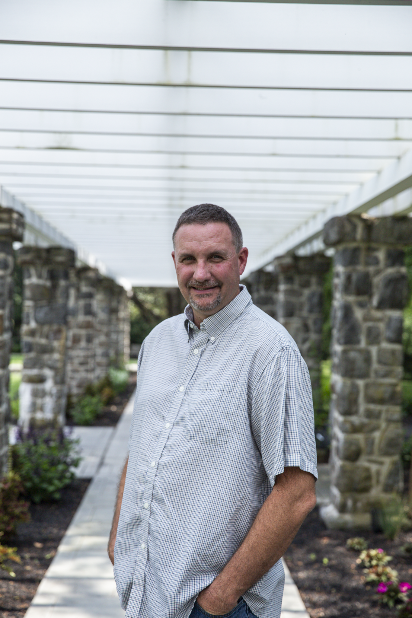 A middle-aged man with short dark hair, a beard, and a mustache standing outdoors under a white pergola on a stone pathway, with gardens on either side.