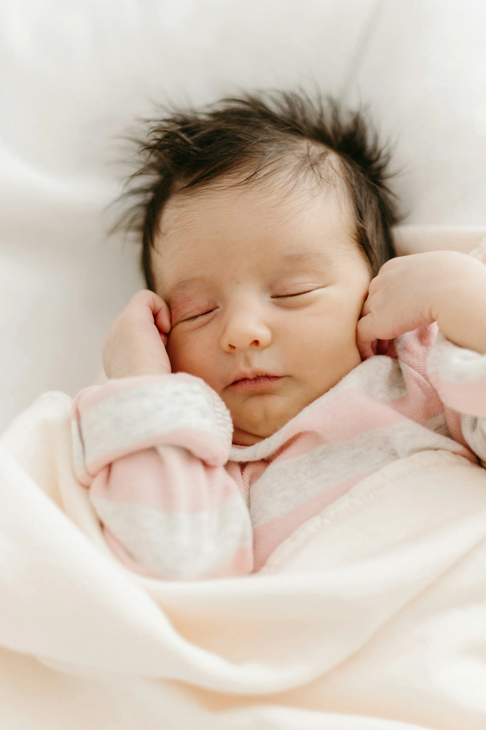A sleeping baby with dark hair, tucked in soft pink and white clothing, resting peacefully with one hand near the face, in a cozy setting.