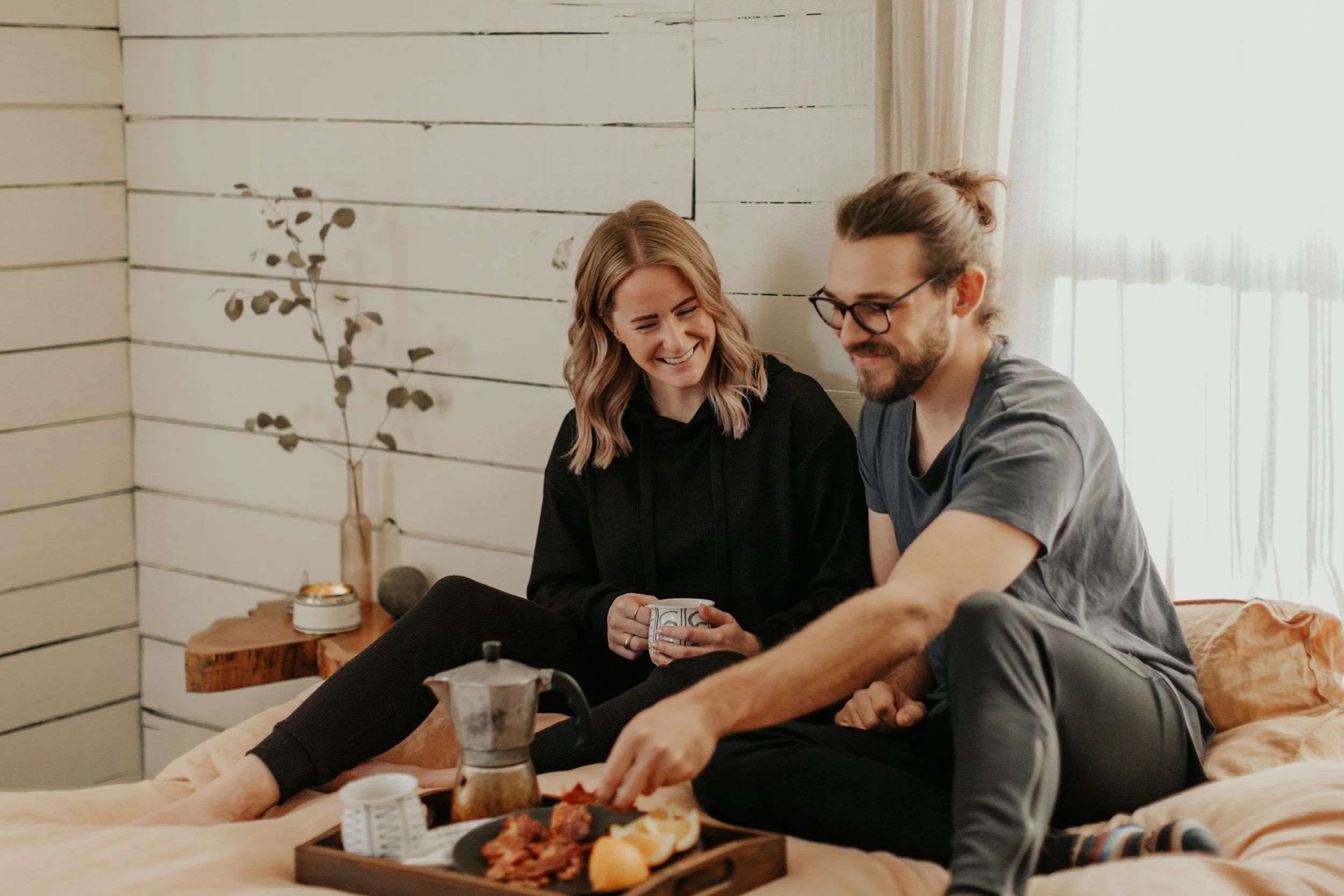 A woman and a man sitting together on a bed in a cozy room, smiling and looking at a smartphone. There is a table with food and drinks in front of them, and a white wooden wall and a window with sheer curtains in the background.