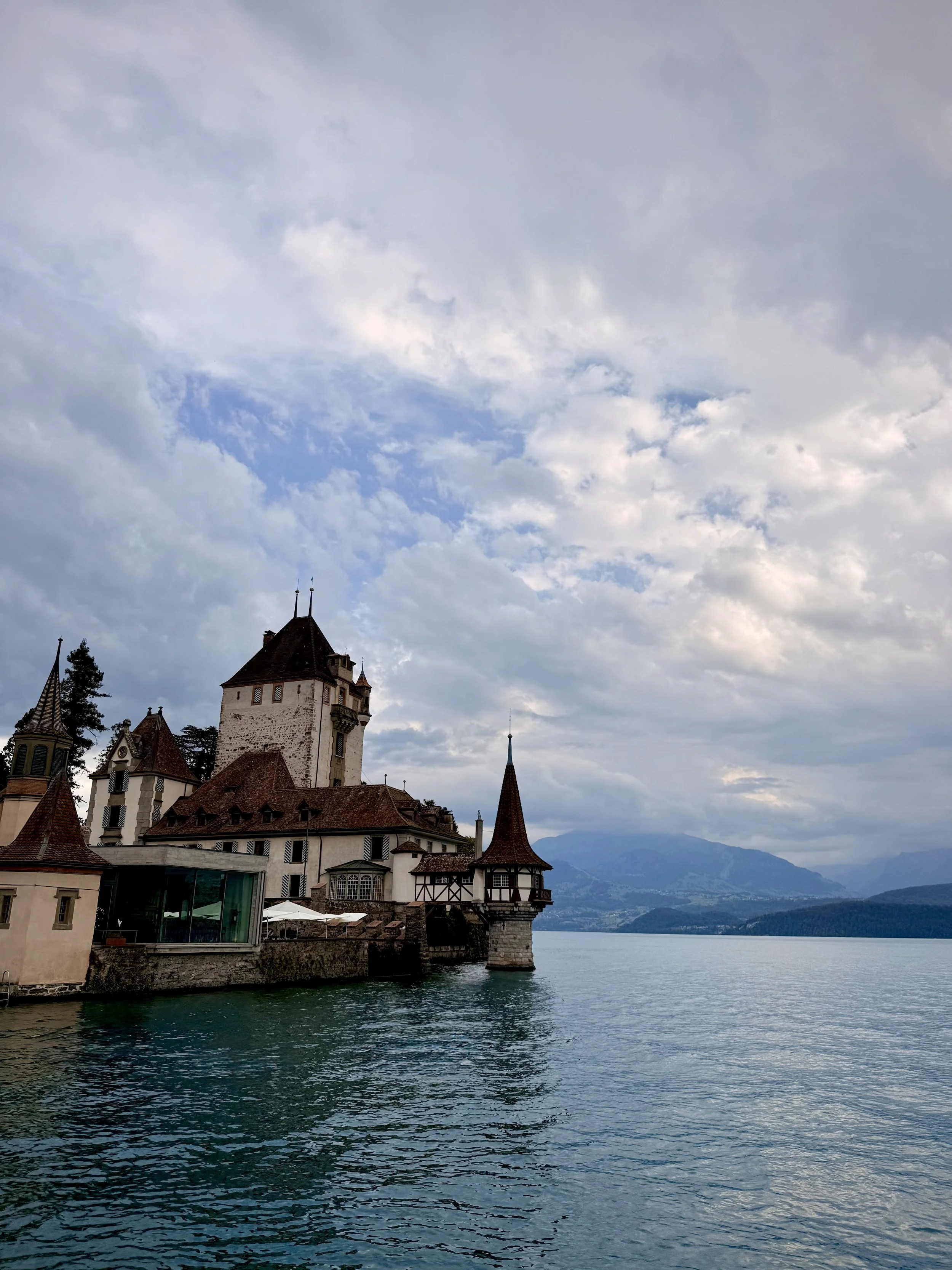 A castle with turrets and towers by a lake, with mountains in the background under a partly cloudy sky.