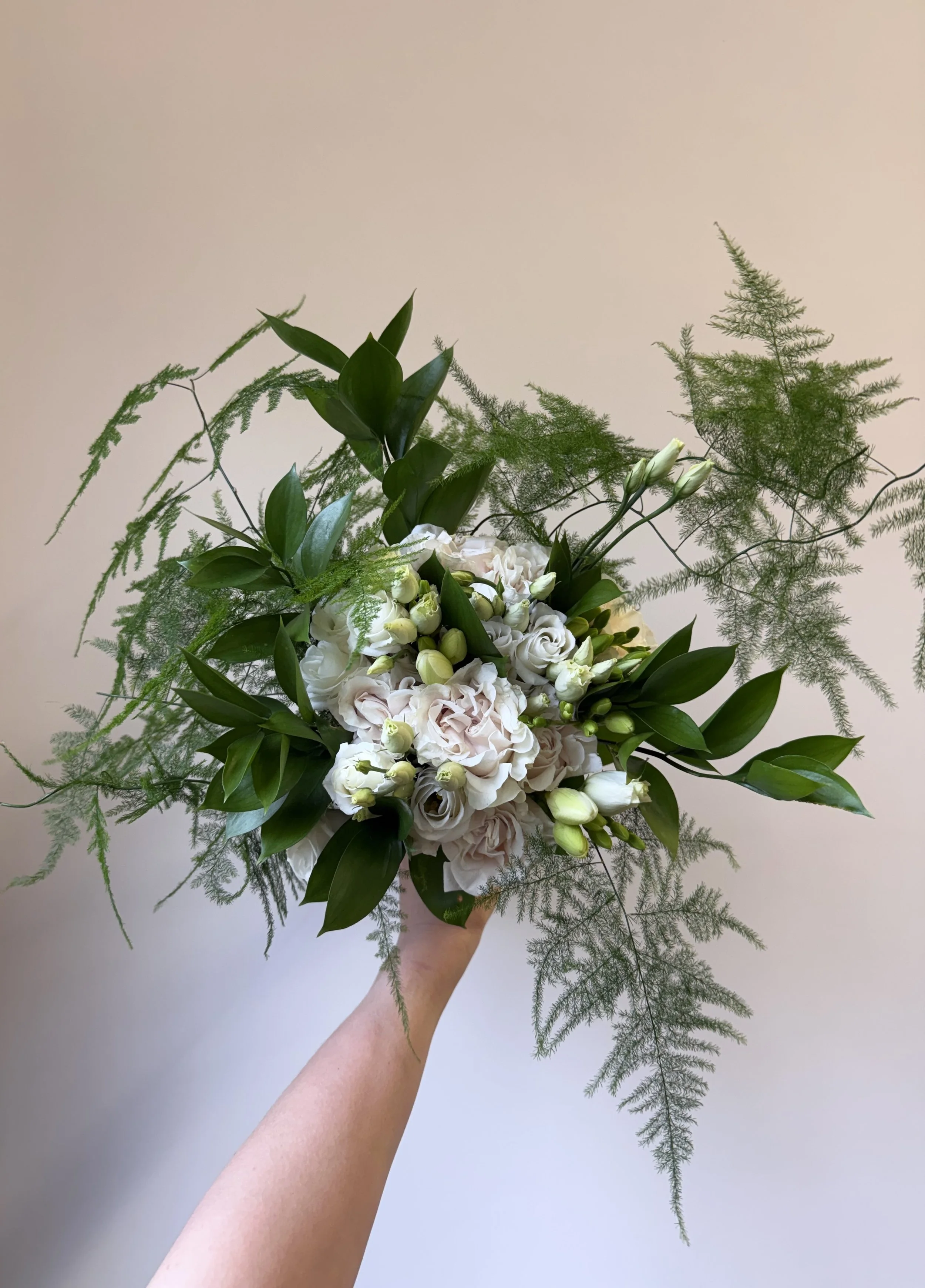 A hand holding a large bouquet of white and green flowers, including roses and greenery, against a plain white wall.