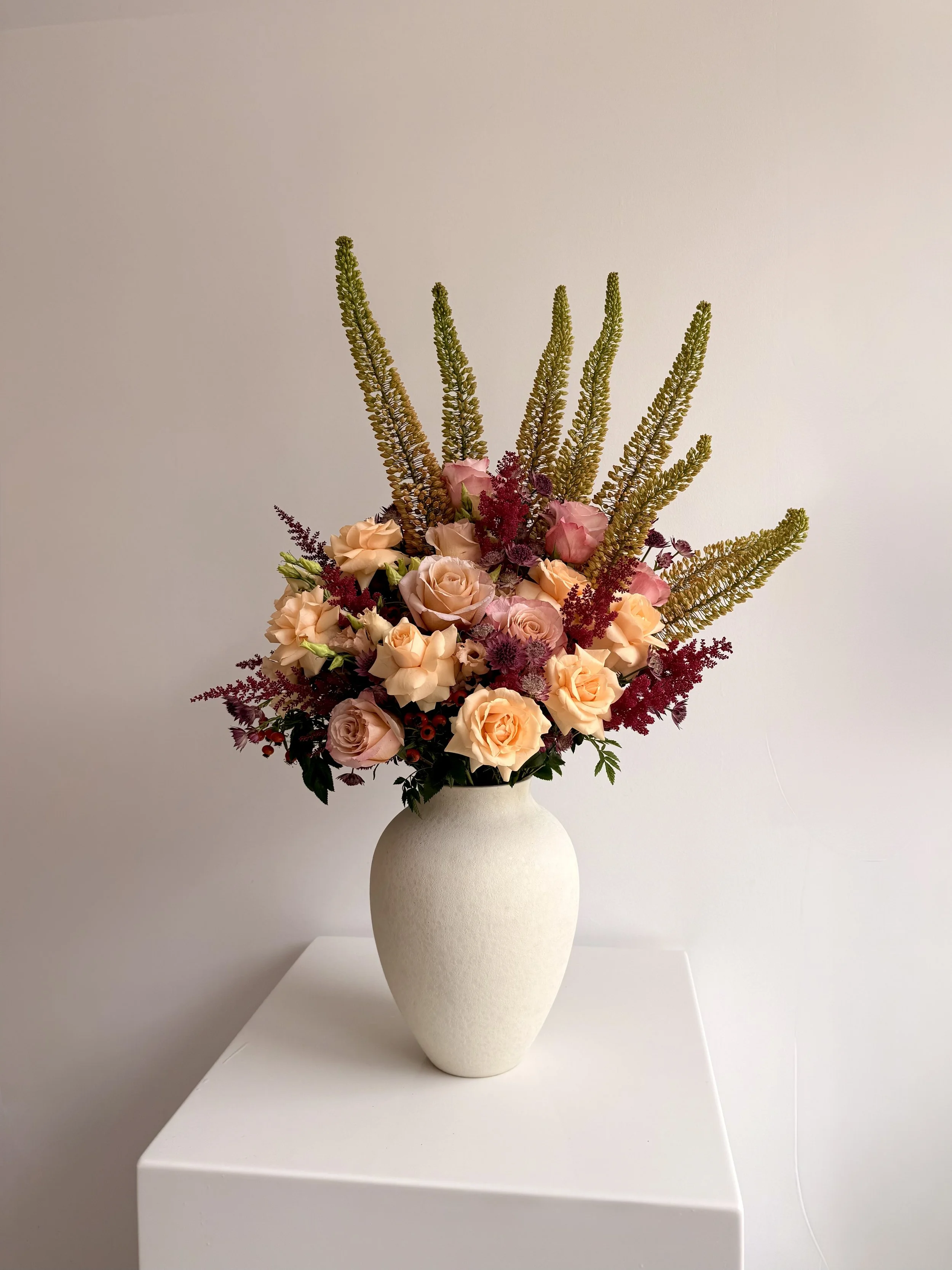 A white ceramic vase holding a large bouquet of peach roses, purple and red foliage, and tall green stems on a white surface against a plain white wall.