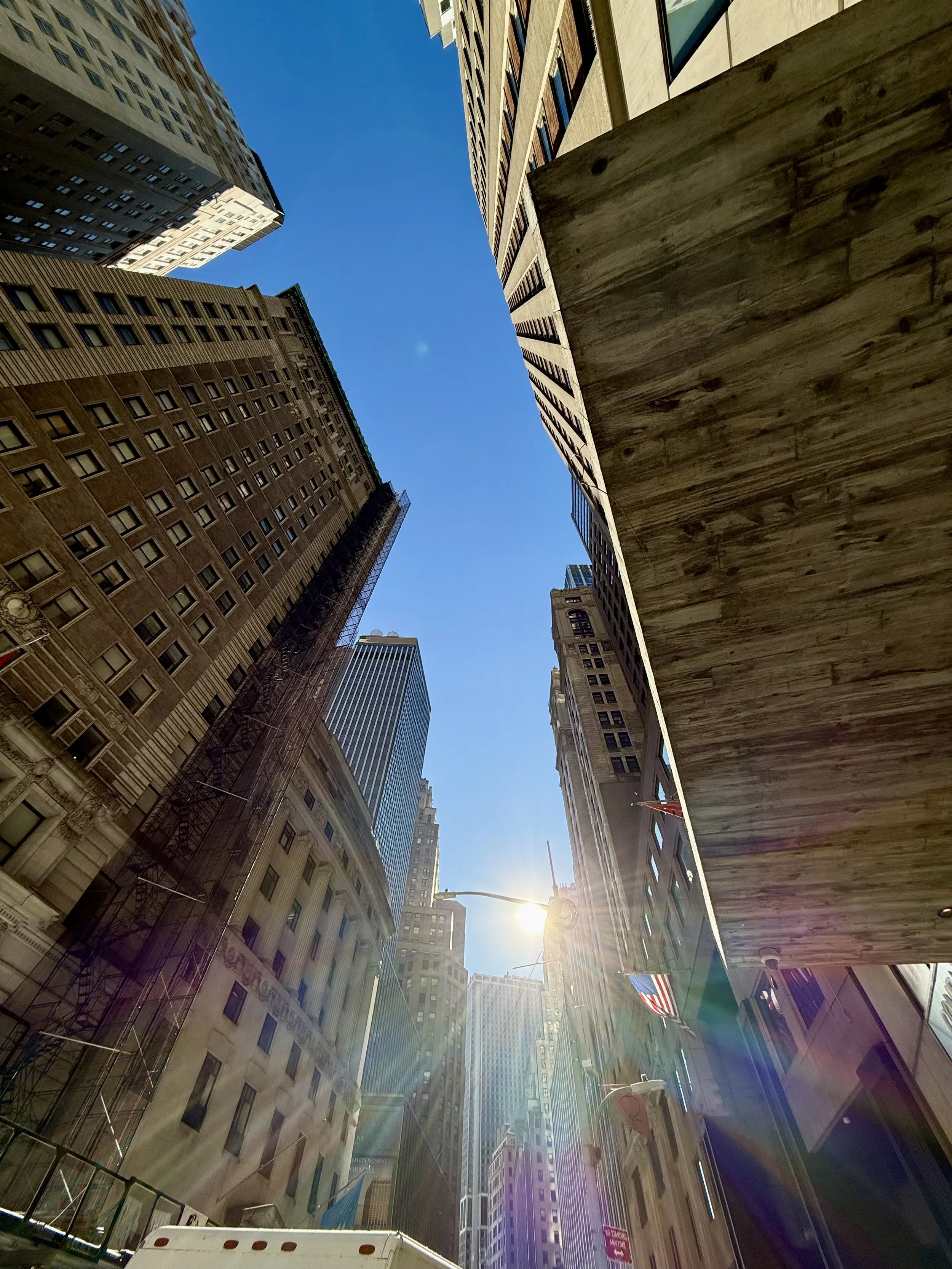Tall buildings in a cityscape with the sun shining between them and a clear blue sky.