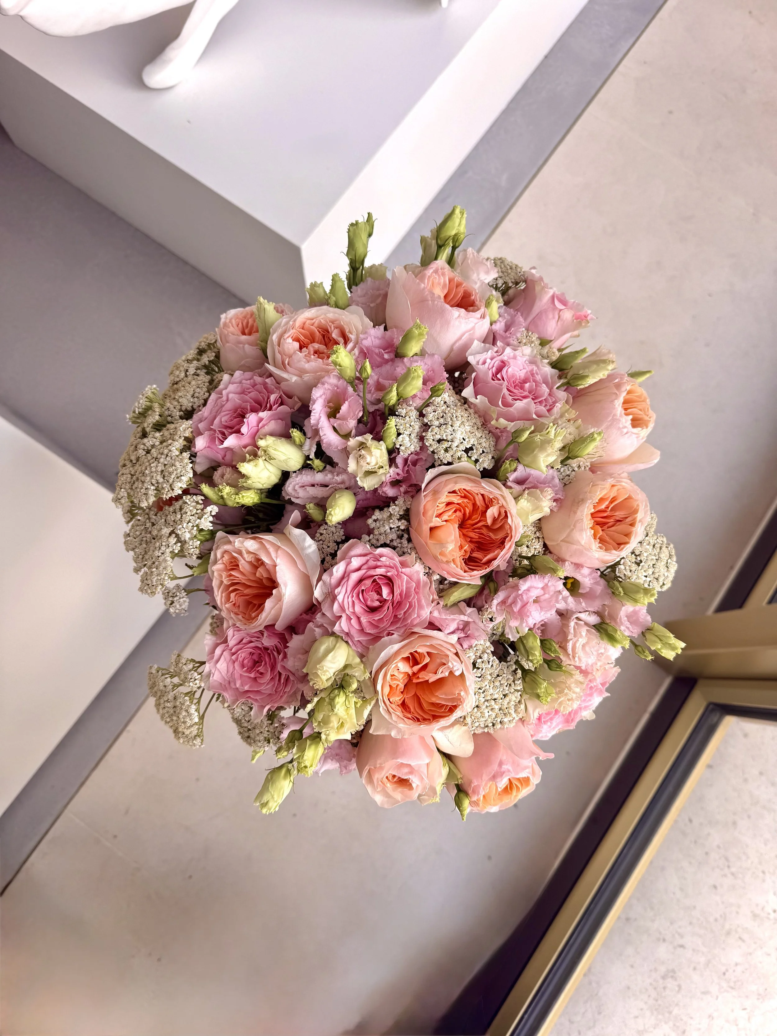 Close-up view of a pink and peach flower bouquet with various roses, lisianthus, and baby's breath, held in front of a gray and white background.