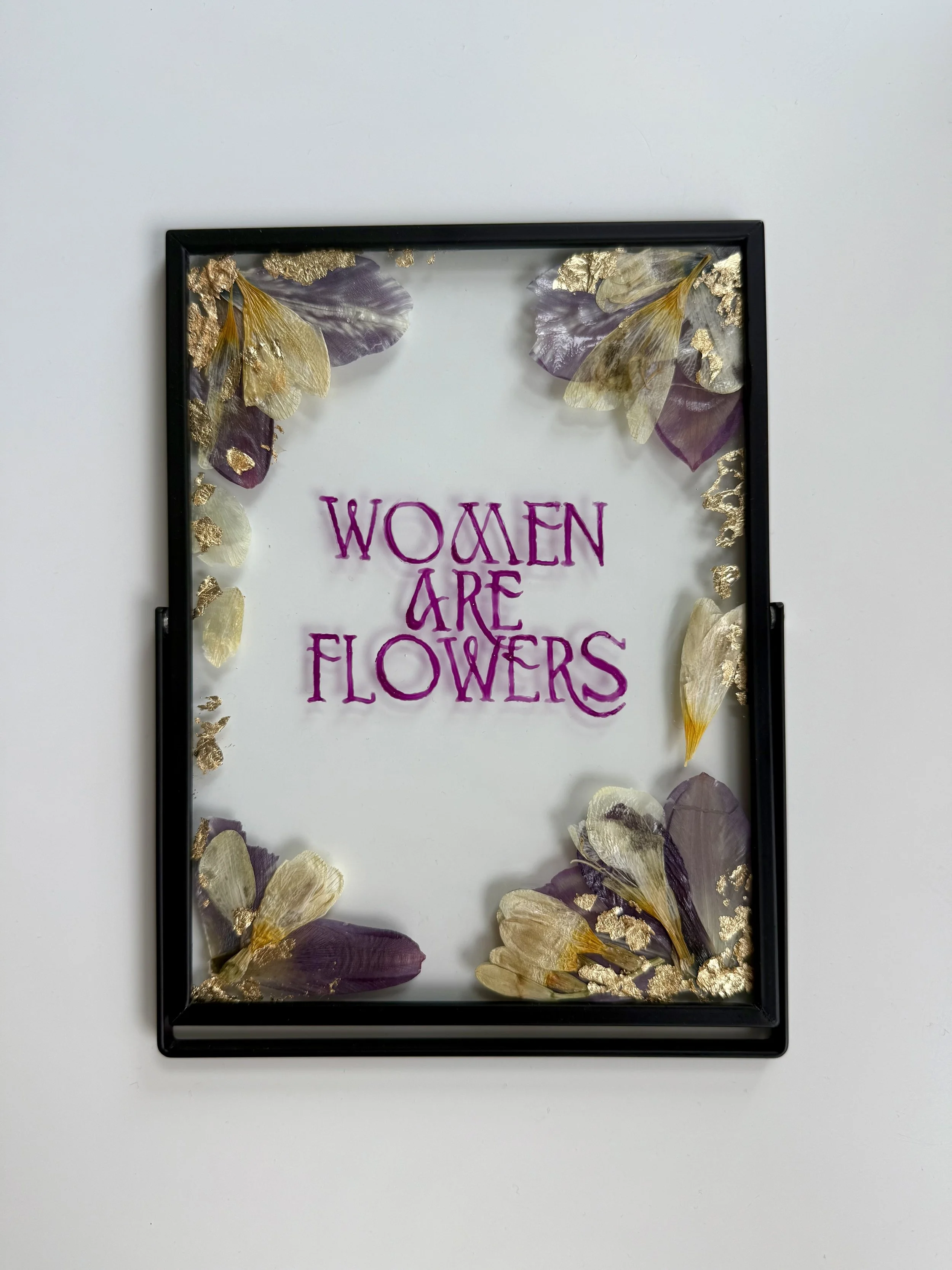 A framed decorative piece with purple and cream-colored butterfly wings and gold leaf accents, featuring the words "Women Are Flowers" written in purple script.