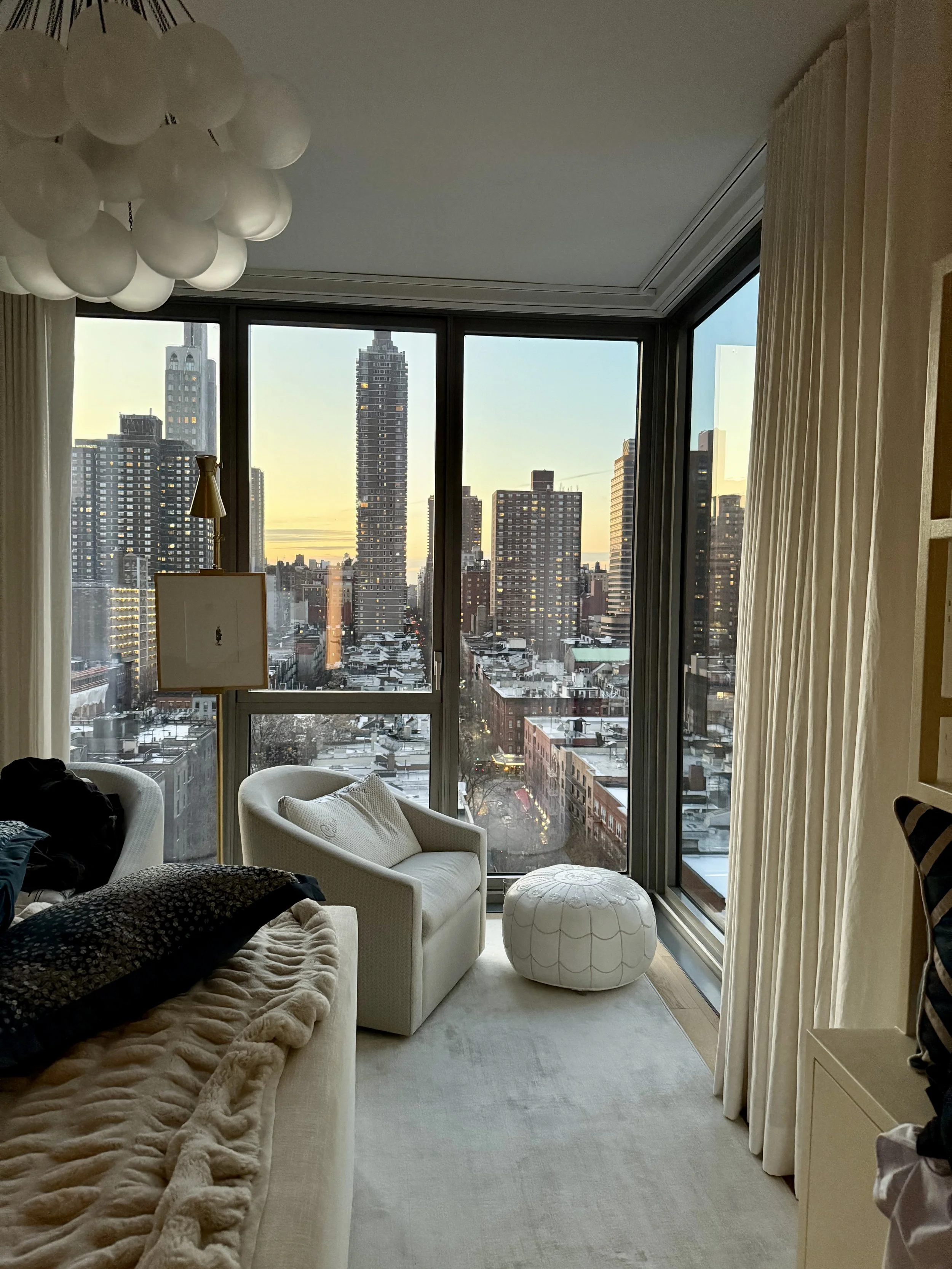 Living room with a white couch, a plush blanket, and black pillows, facing large floor-to-ceiling windows showing a city skyline at sunset, with high-rise buildings, some with lights on, and snow-covered rooftops, and a large white pouf in front of t