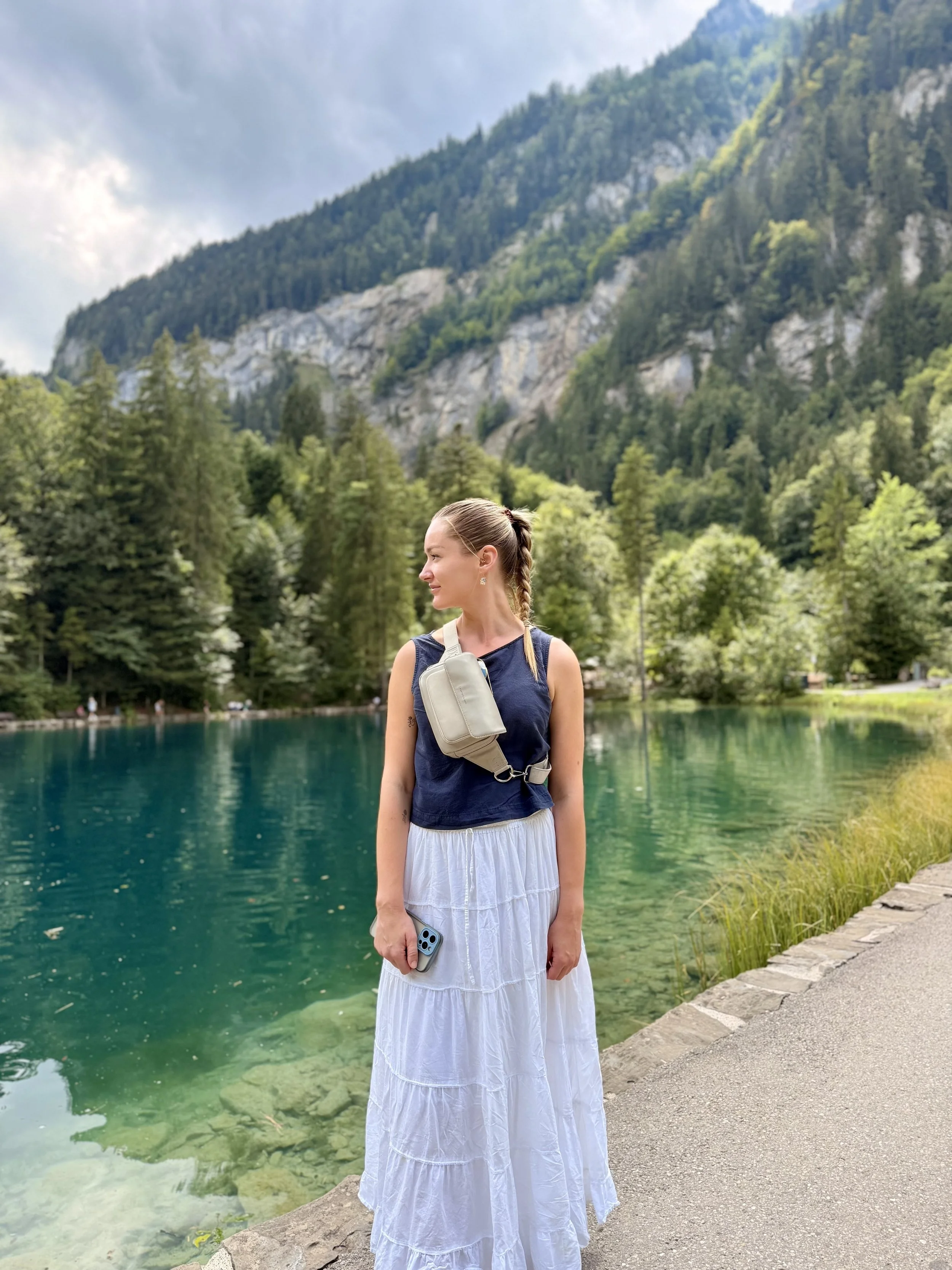 A woman with braided hair standing by a green lake surrounded by forest and mountains, holding a mobile phone and wearing a light-colored crossbody bag.