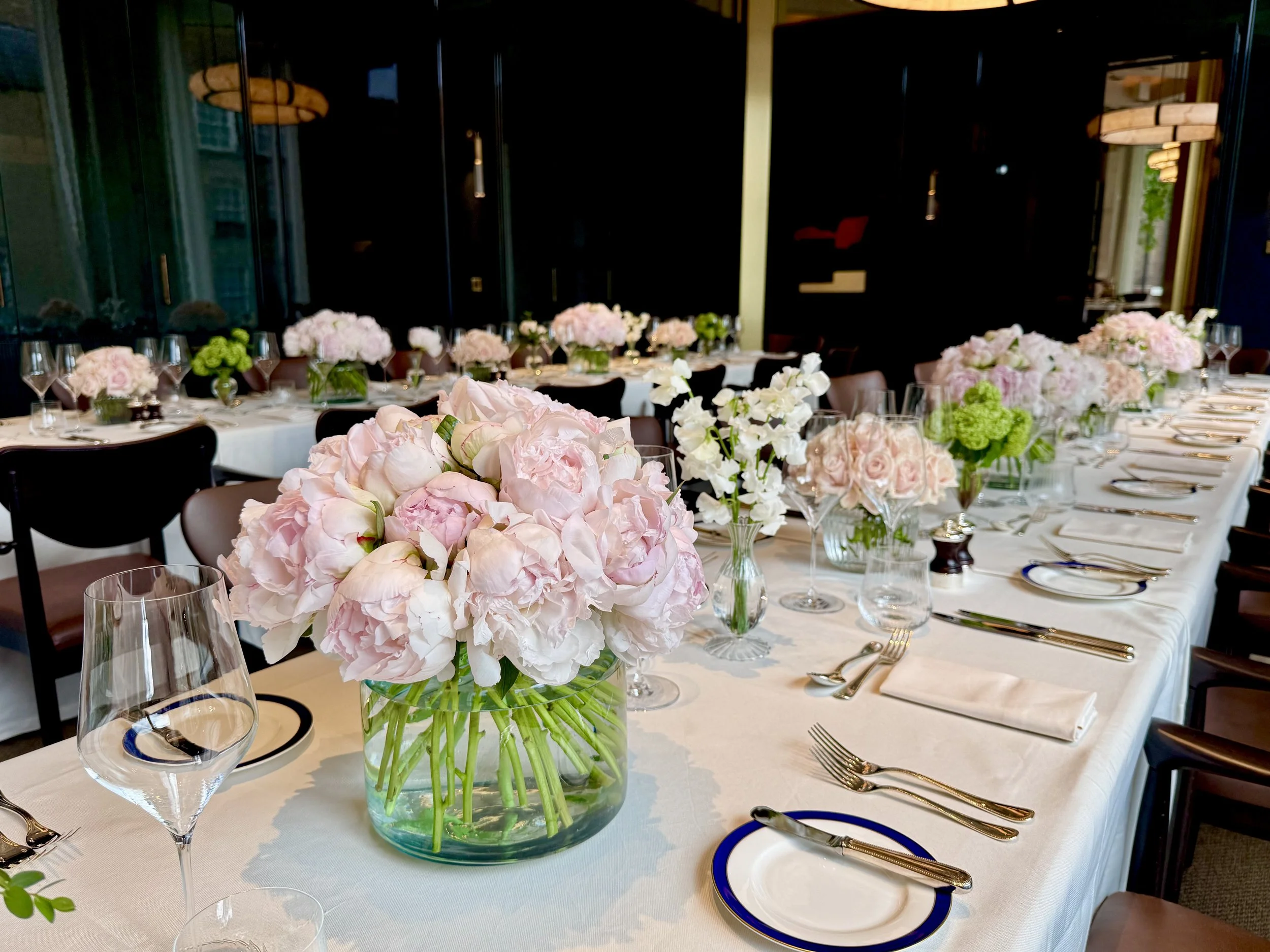 A banquet table decorated with pink and white floral centerpieces, set with white plates, silver cutlery, and wine glasses in a well-lit dining room.