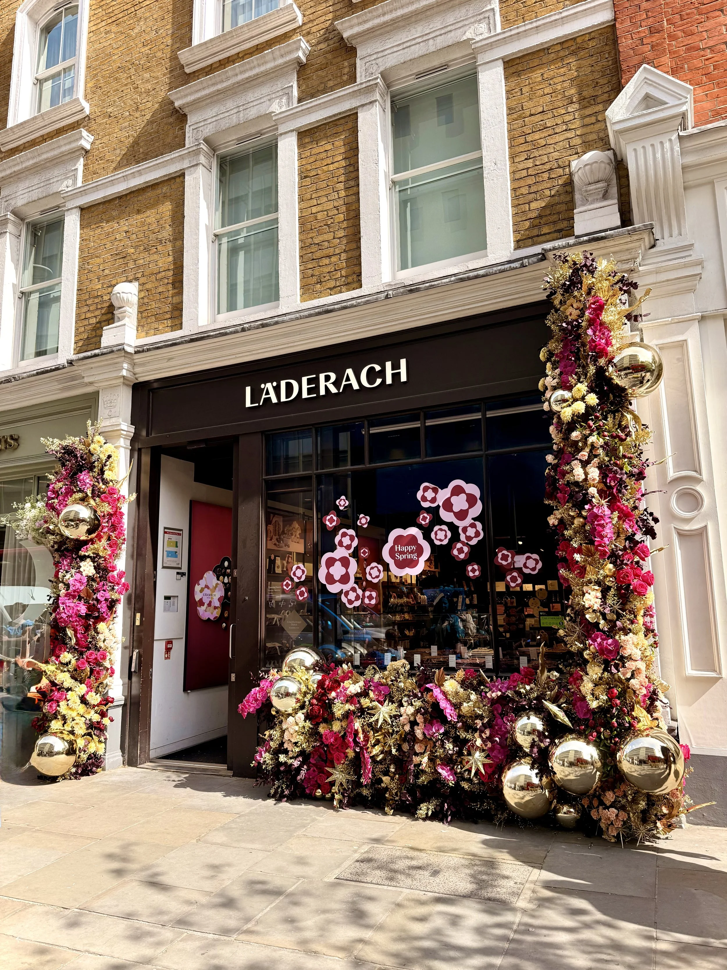 Storefront with the sign 'Läderach' decorated with floral arrangements and golden ornaments for spring, with a brick building above.