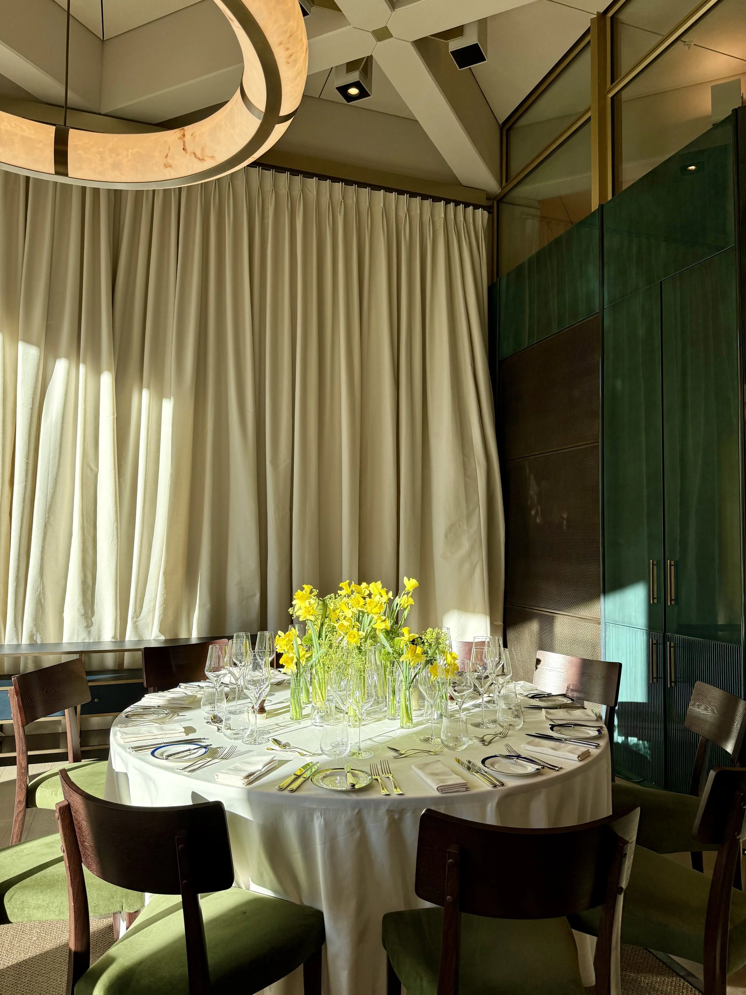 Round dining table with white tablecloth, set with silverware, glassware, and white napkins, decorated with a yellow flower centerpiece, surrounded by wooden chairs with green upholstered seats, in a room with cream curtain and green cabinet.