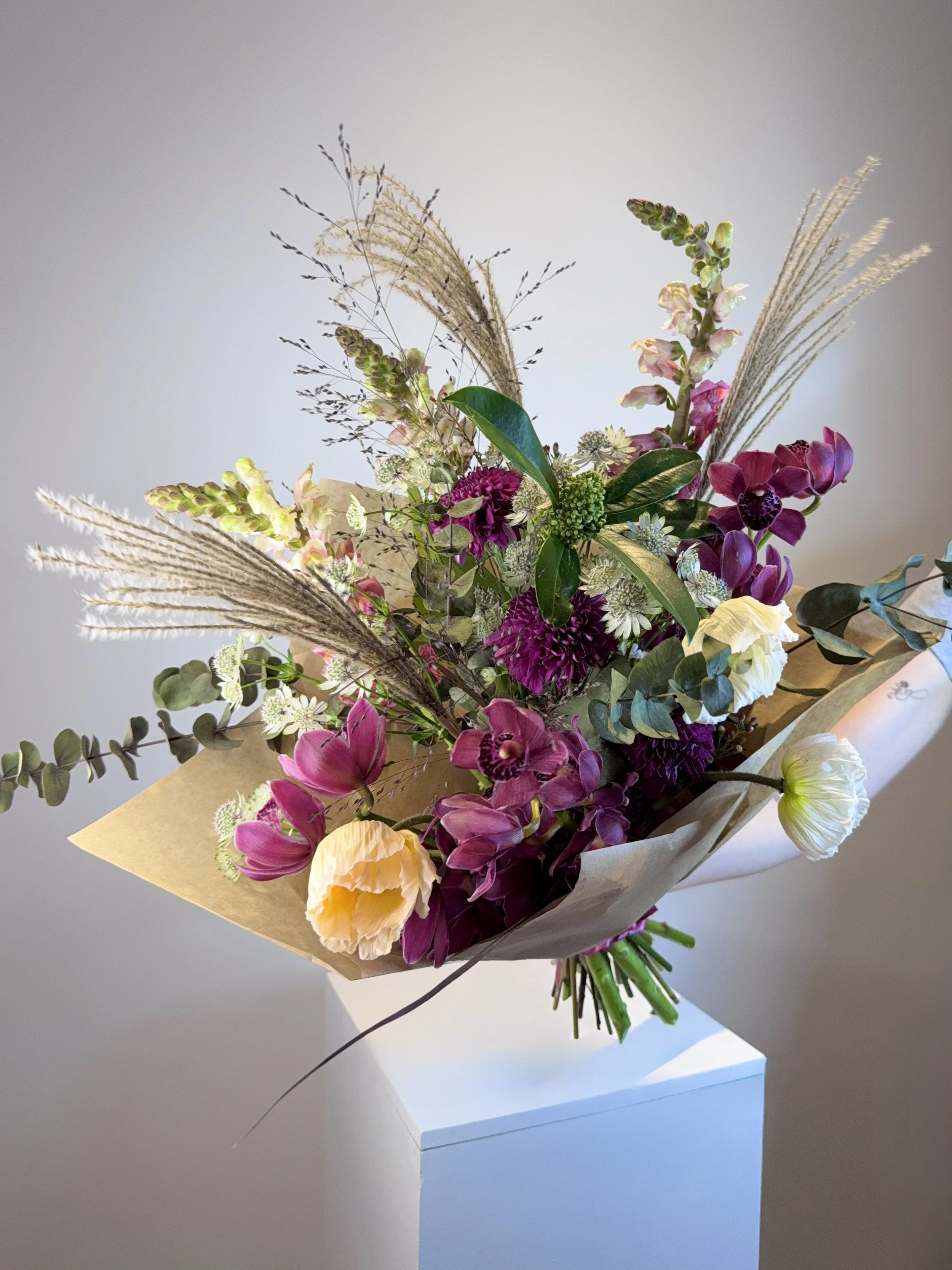 A bouquet of mixed flowers including pink orchids, white and yellow roses, and various greenery, wrapped in beige paper, placed on a white pedestal against a light gray background.