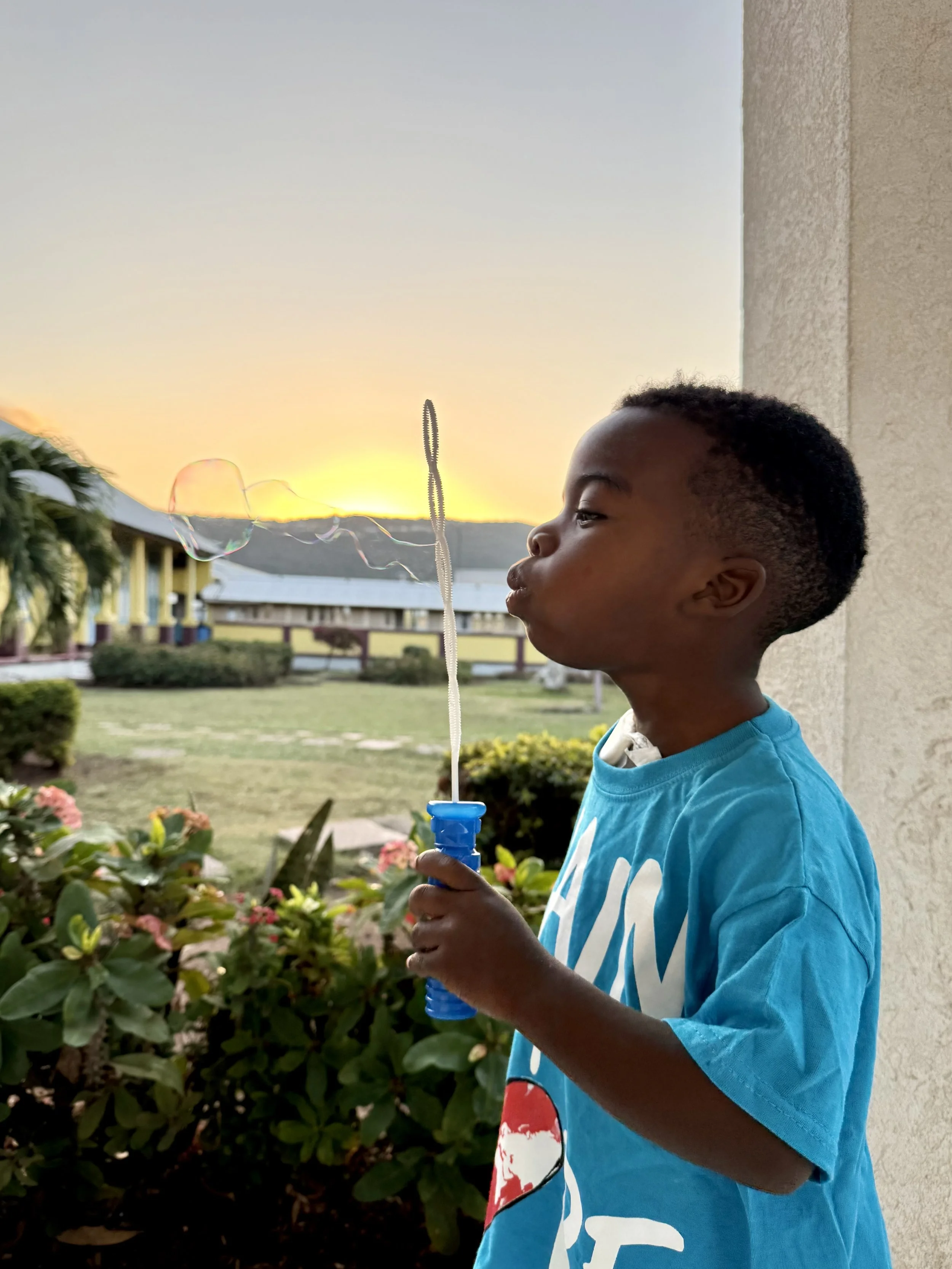 A boy in a blue T-shirt is blowing bubbles using a bubble wand outside during sunset, with a garden, bushes, and apartment buildings in the background.