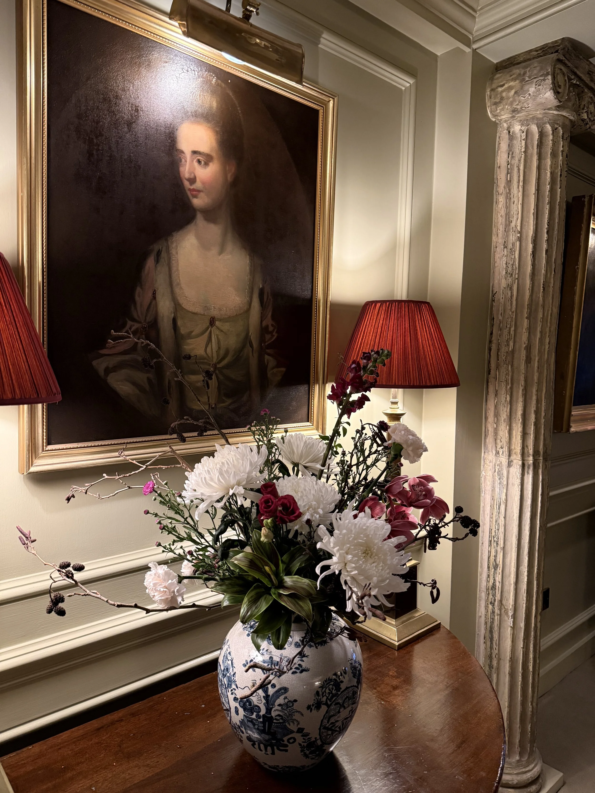 A vase with white, pink, and purple flowers on a wooden table in front of a radiator and wall, with a portrait painting hanging on the wall beside a column.