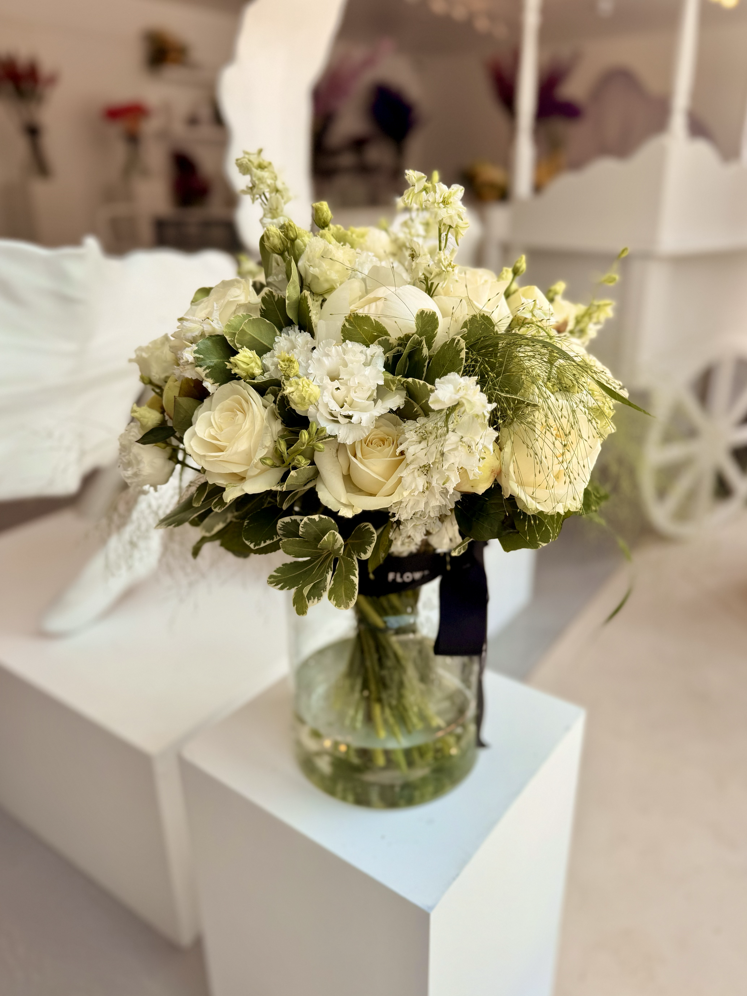 A bouquet of white roses, carnations, and other white flowers with green foliage arranged in a glass vase placed on a white table.