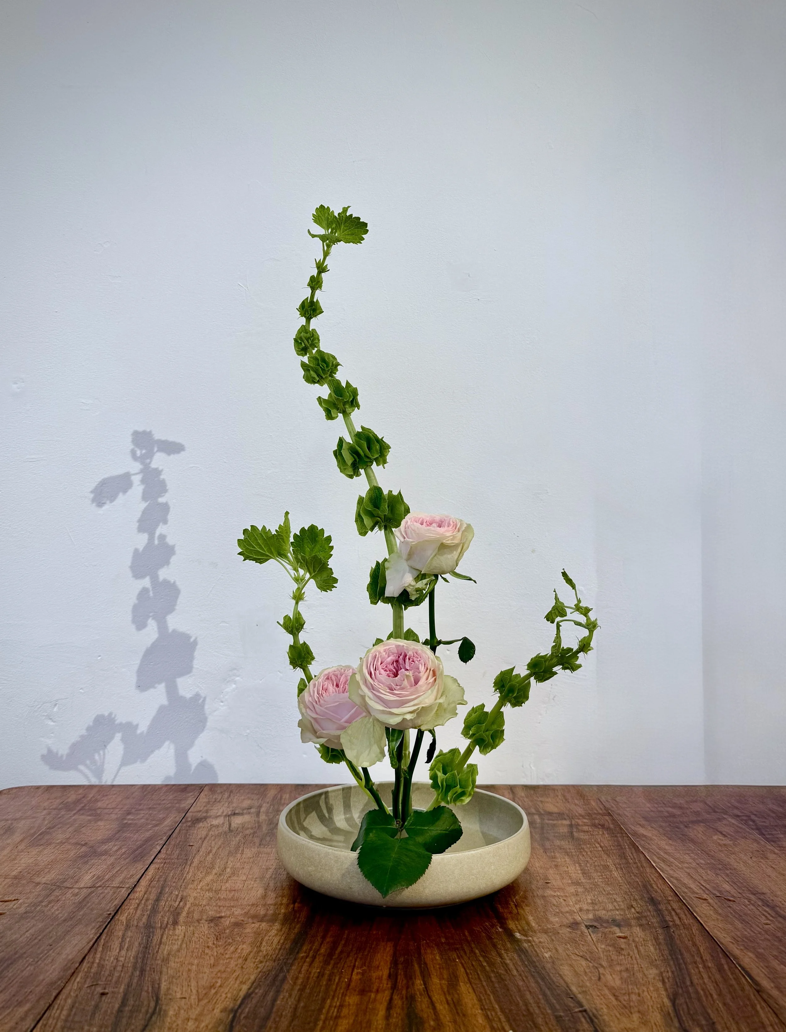 A tall flower arrangement with pale pink roses and green leafy stems is placed in a round beige ceramic bowl on a wooden table against a plain white wall.
