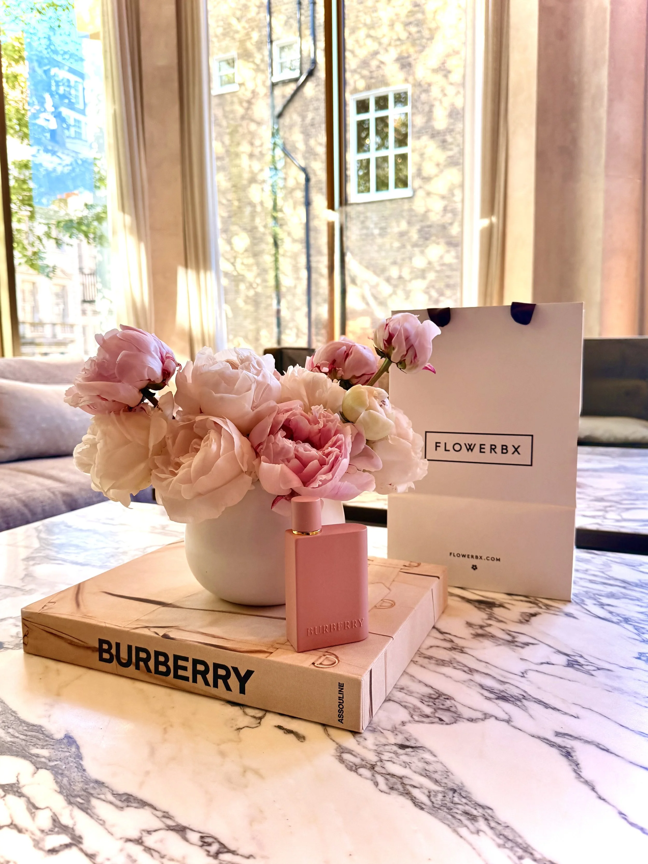 A bouquet of pink and white flowers in a white vase, sitting on a wooden box with the Burberry logo, on a marble table. There is a white FlowerBX box and a pink Burberry perfume bottle nearby. The background shows large windows with curtains and a br