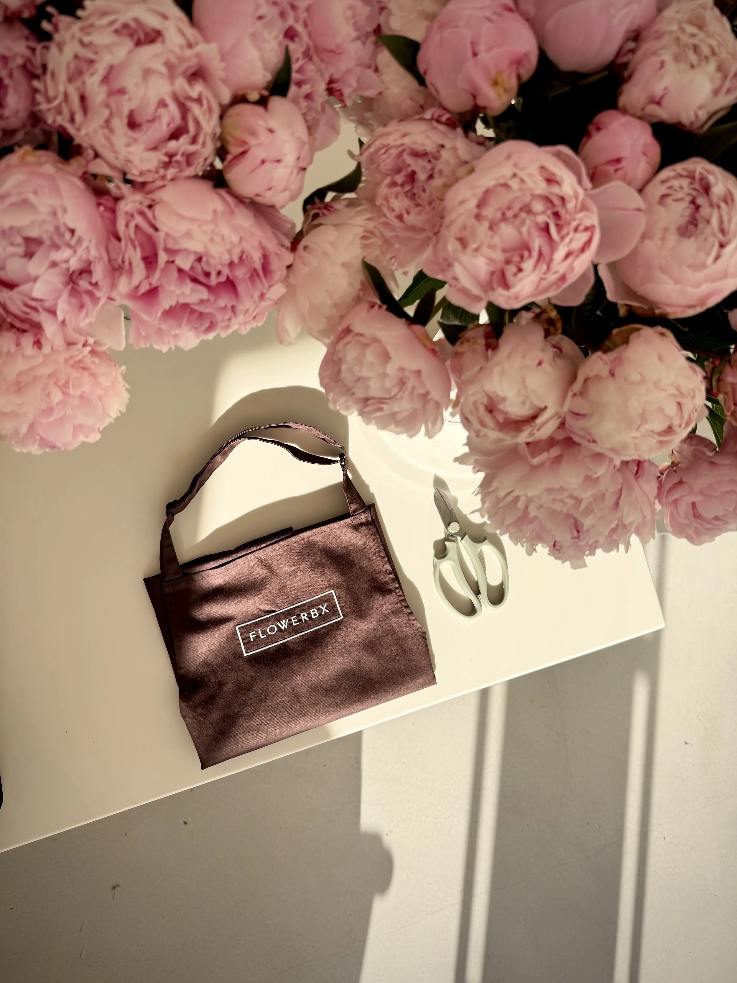 A bouquet of pink peonies on a white surface, a pair of scissors, and a brown SatinFlowerX shopping bag.