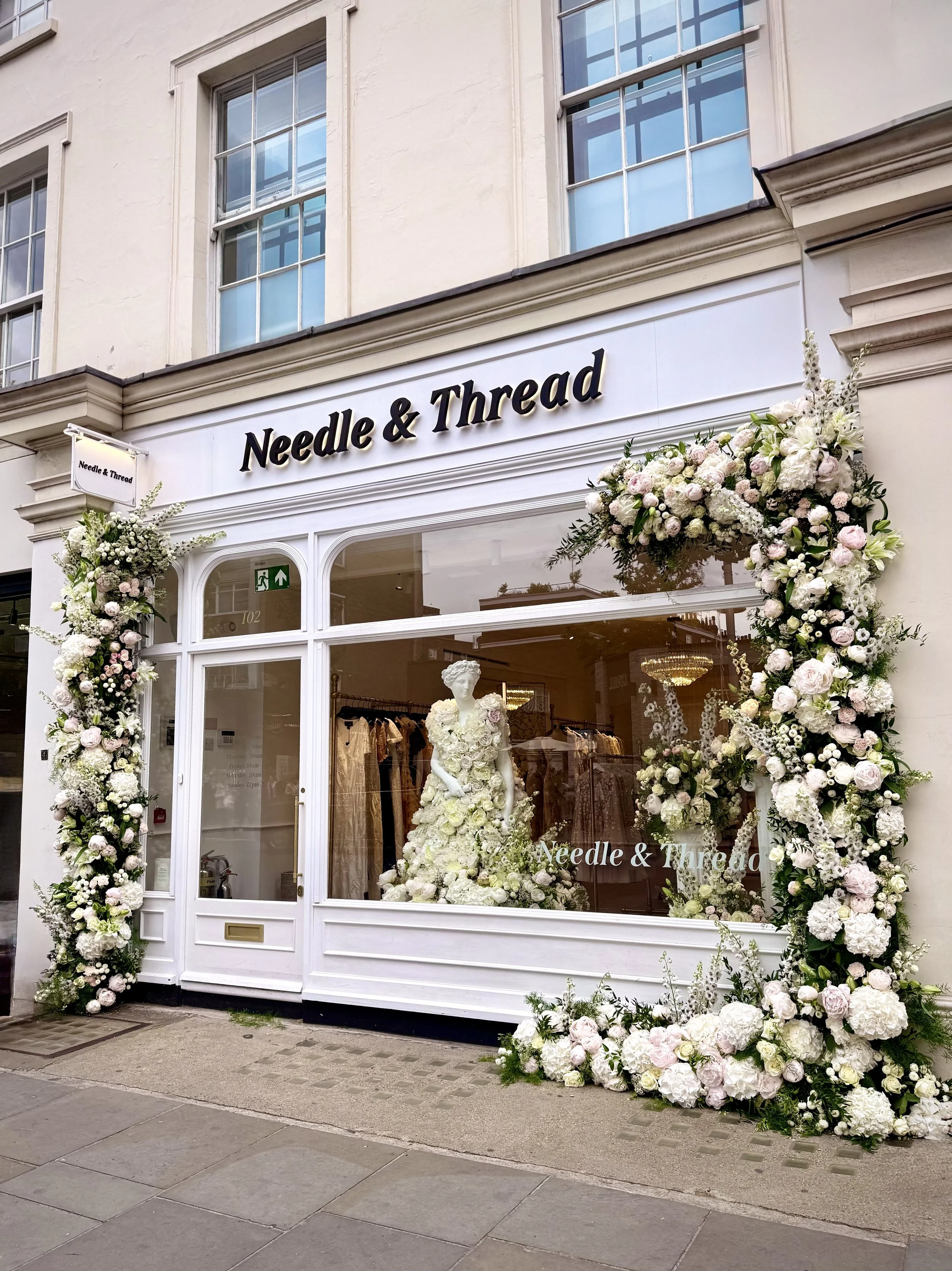 The storefront of 'Needle & Thread,' a clothing store, decorated with large floral arrangements of white and pale pink flowers at the entrance.
