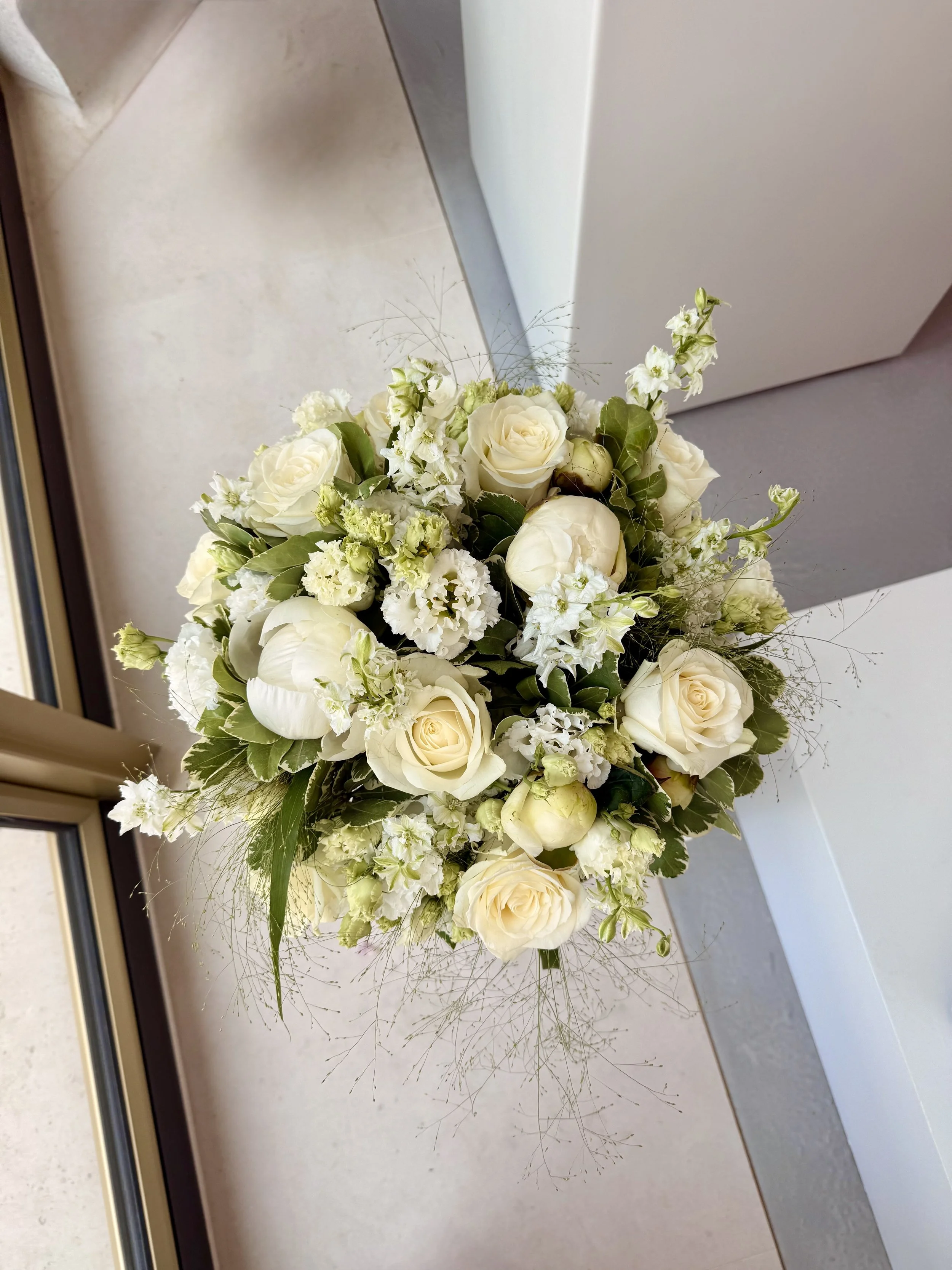 A close-up of a bouquet of white roses and other white flowers with green leaves and sprigs of greenery.