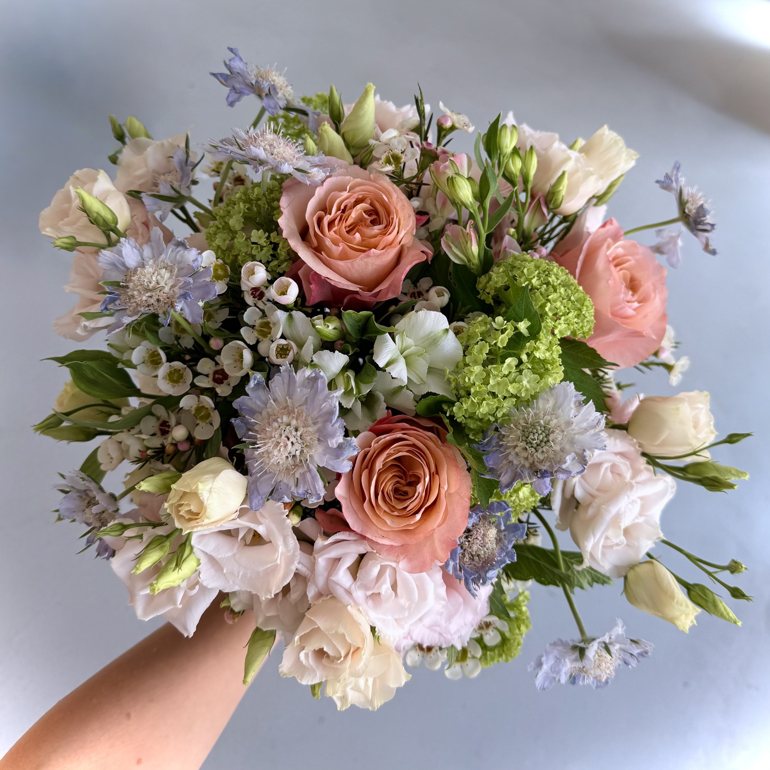 A hand holding a bouquet of various pastel-colored flowers, including roses, hydrangeas, and other small blossoms, against a plain light background.