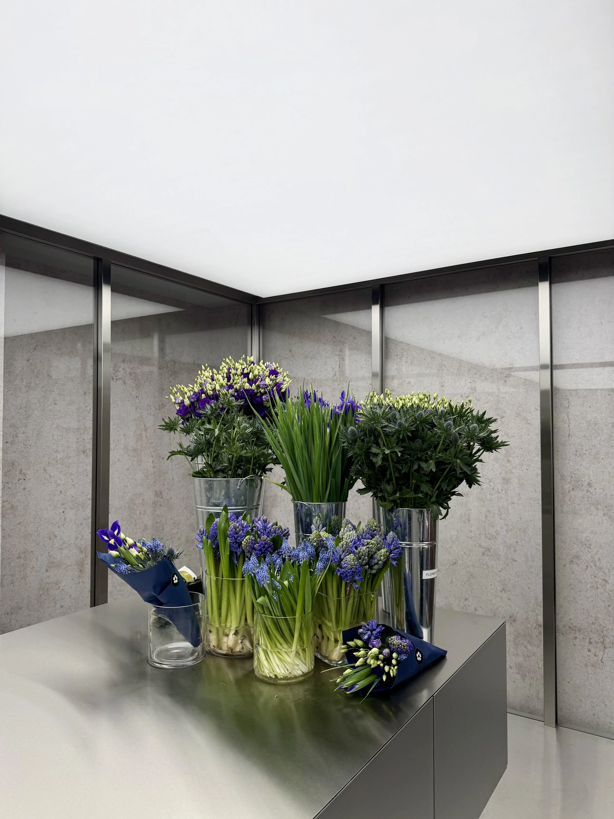 Various flower arrangements in vases on a gray table in a modern, minimalistic room with light walls and glass panels.
