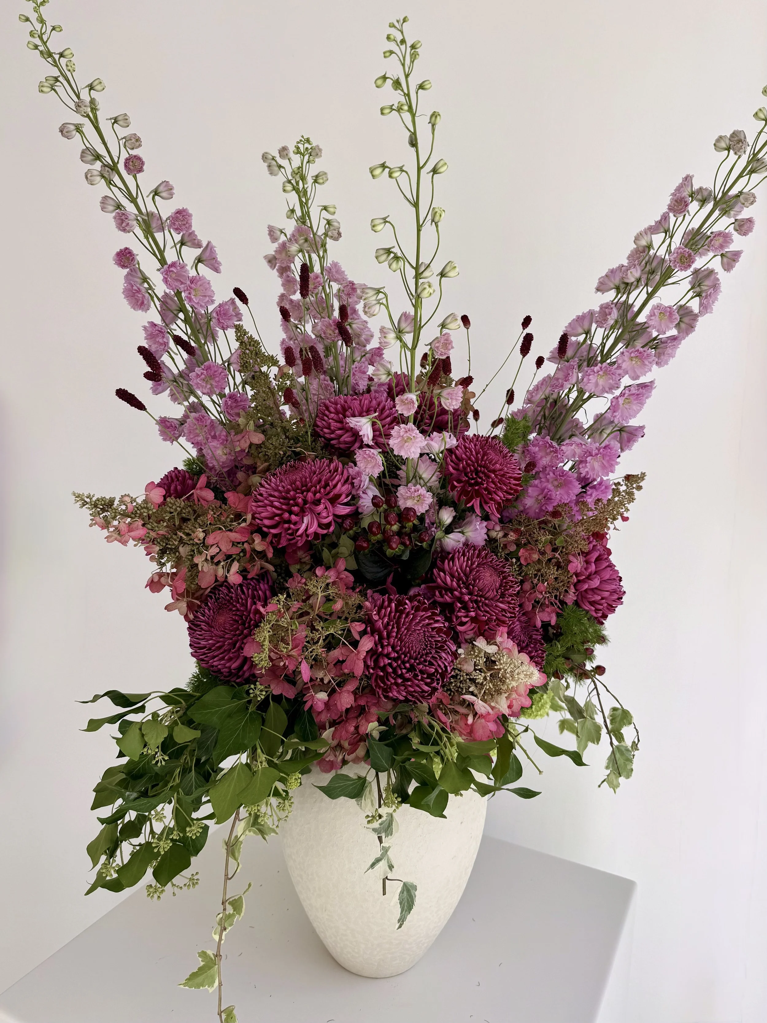 A tall white vase containing a colorful bouquet of pink and purple flowers with green leaves and sprigs, arranged on a white surface against a plain white background.