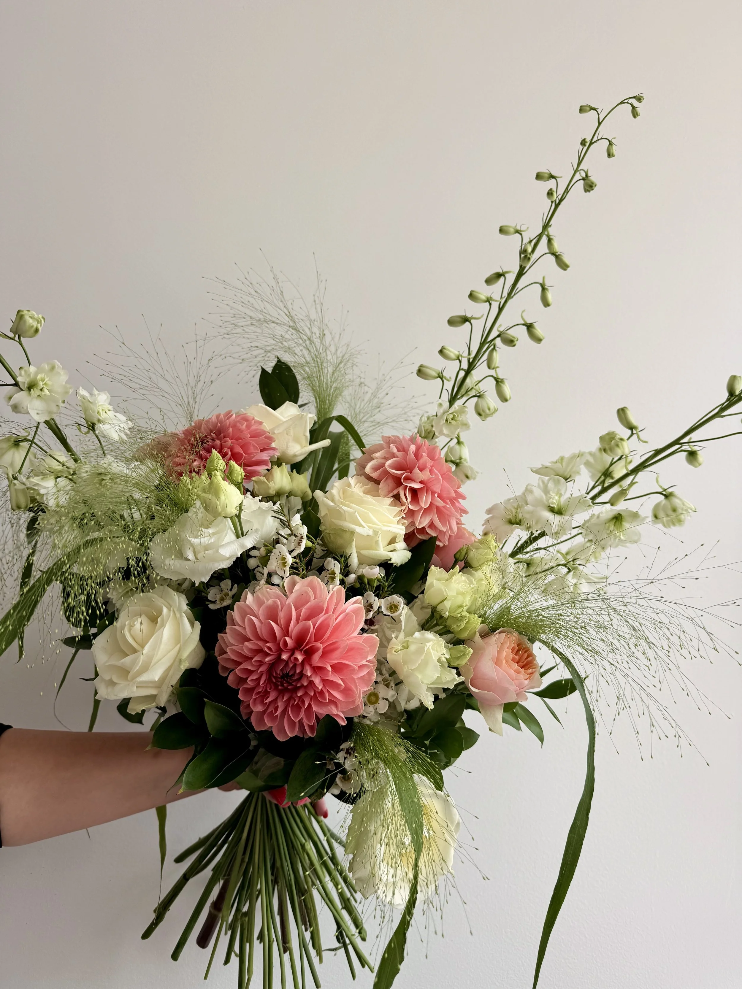 A bouquet of mixed flowers including pink dahlias, white roses, and various green foliage, held by a person's hand against a plain light-colored background.