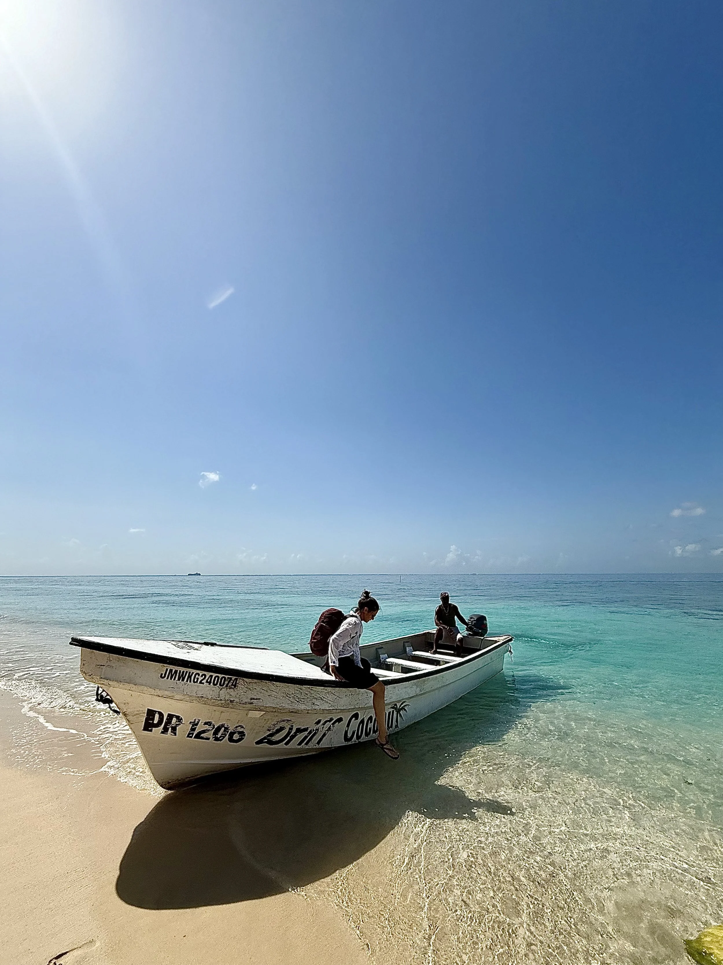A small motorboat with two people on a sandy beach, one sitting in the boat and the other standing on the shore, under a bright blue sky with a few clouds and calm ocean waters in the background.