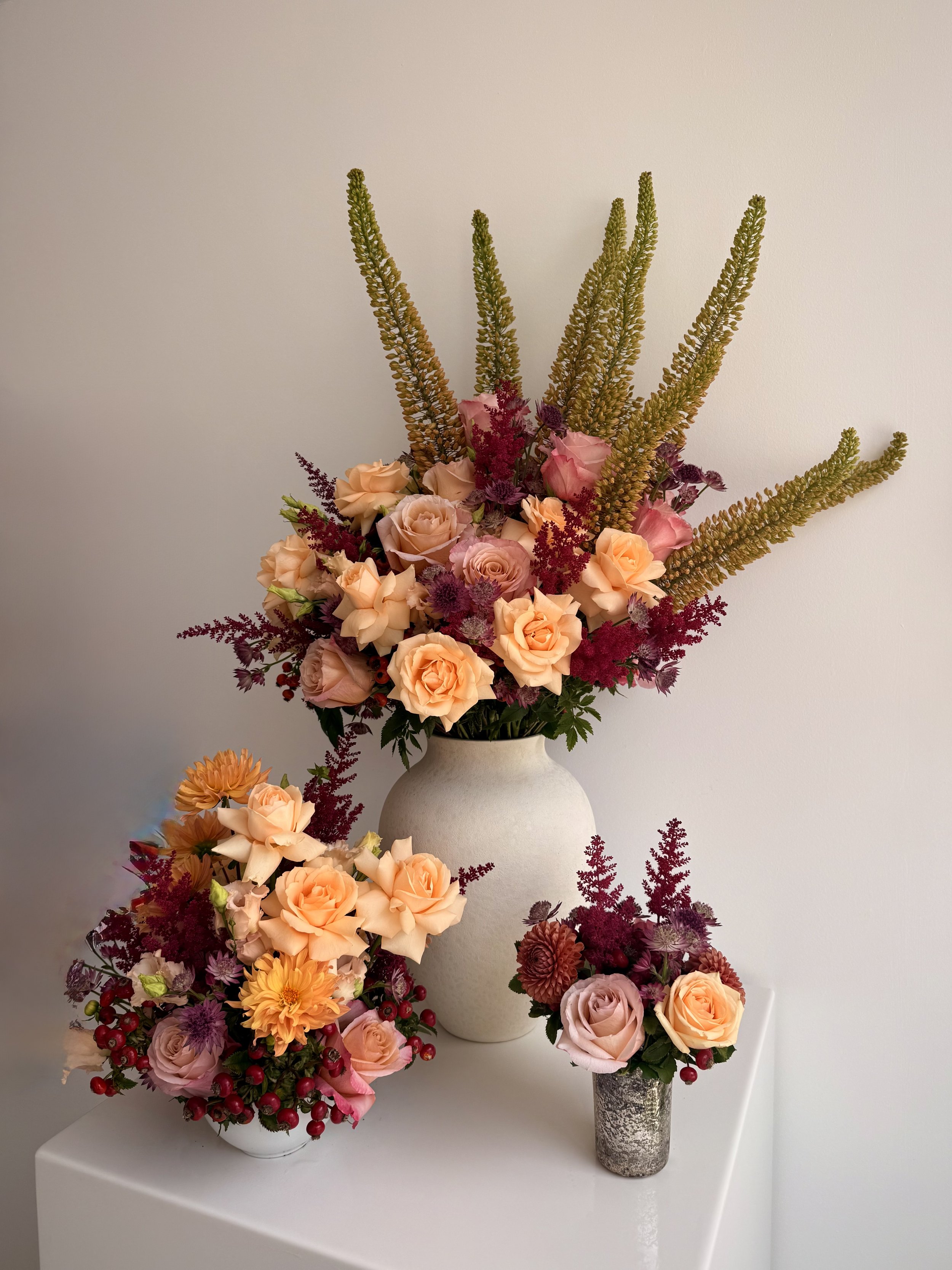 A white vase holding a colorful floral arrangement with pink roses, peach roses, purple accents, and green foliage, placed on a white surface against a plain white wall.
