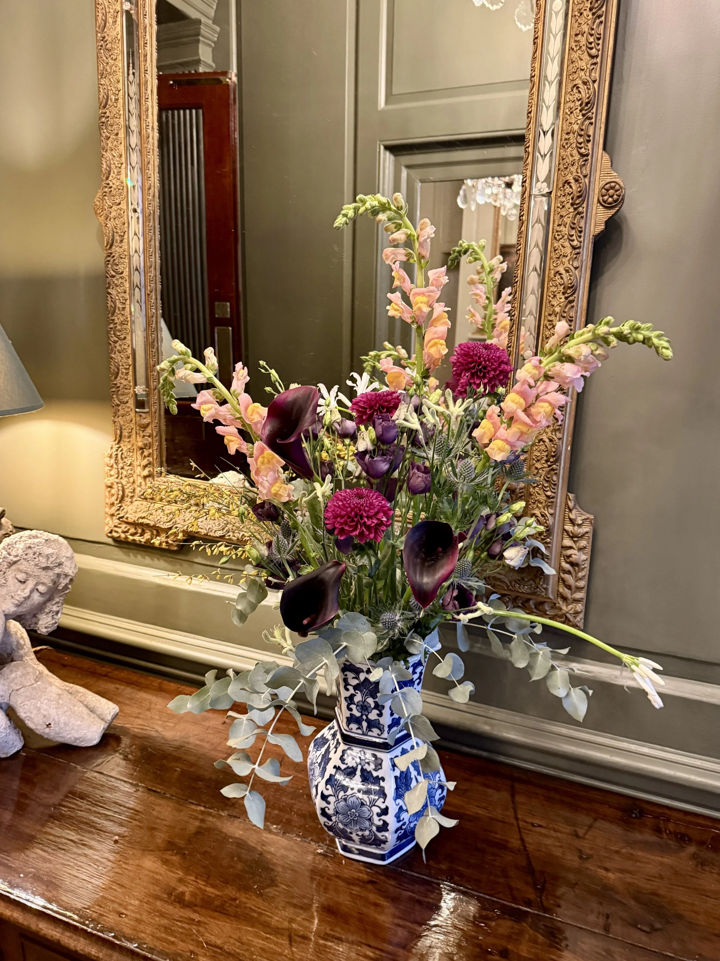 A floral arrangement in a blue and white patterned vase, placed on a wooden table, with a large ornate mirror and a chandelier visible in the background.