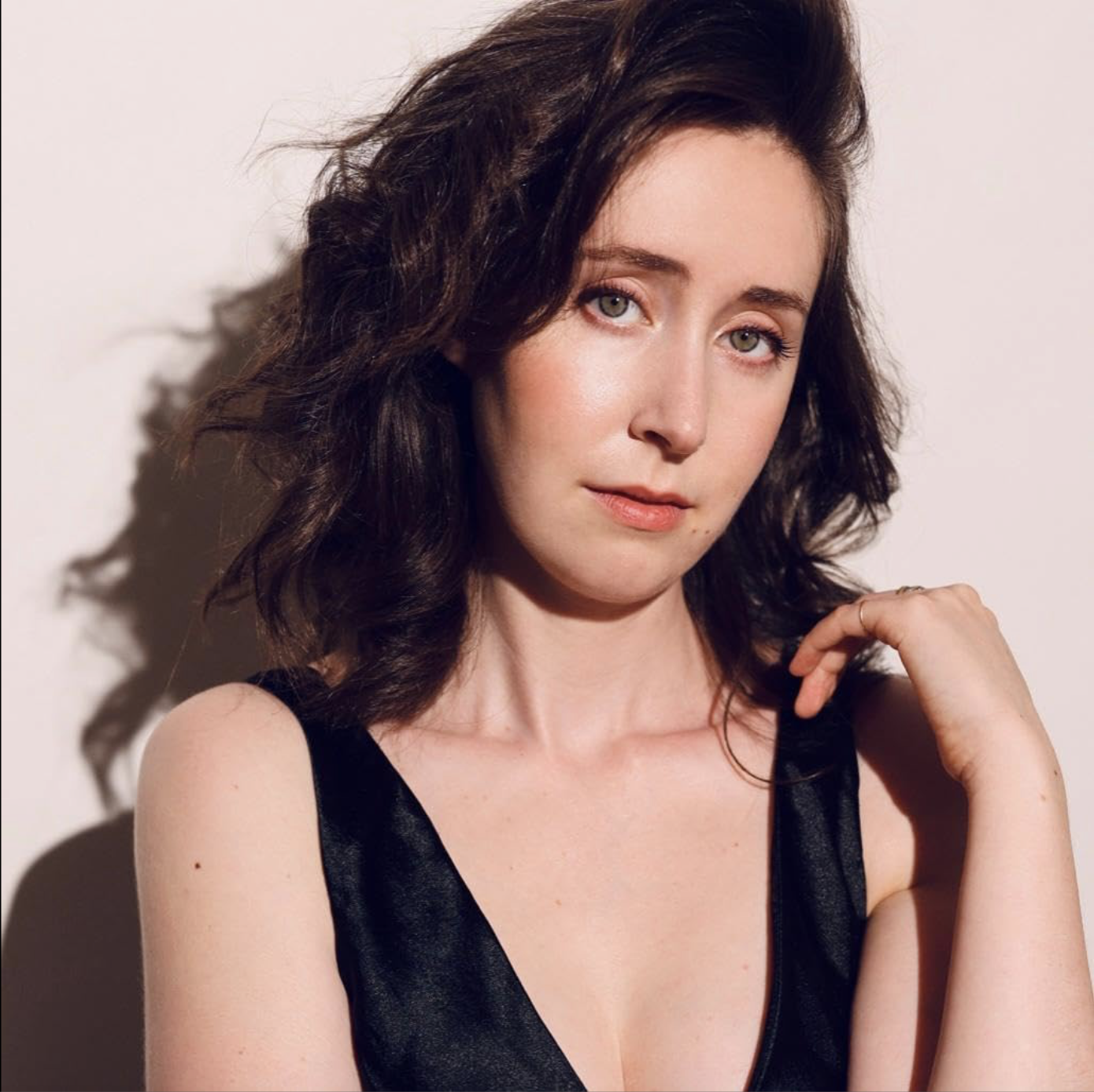 A woman with shoulder-length dark curly hair wearing a black sleeveless top, resting her chin on her right hand, looking into the camera with a slight smile, in front of a plain light-colored background.