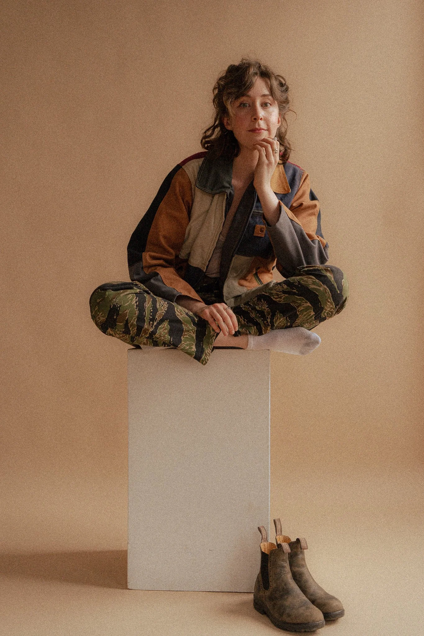 A woman with curly hair sitting cross-legged on a white cube, wearing a jacket and camouflage pants, with a pair of worn boots beside her, against a beige background.