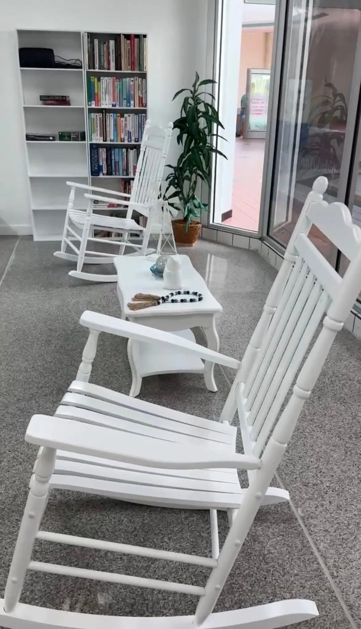 Indoor sitting area with white rocking chairs arranged around a small white table, a tall potted plant, a bookshelf filled with books, and a glass door letting in natural light.