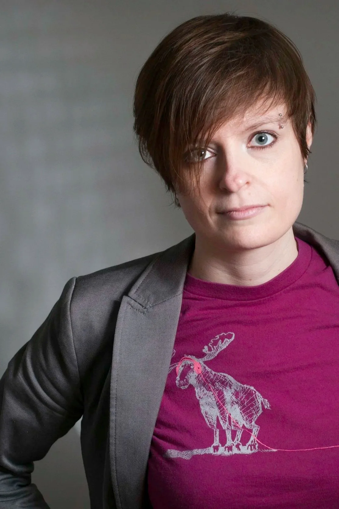 A woman with short brown hair and fair skin wearing a gray blazer over a pink T-shirt with a moose graphic, looking directly at the camera against a plain gray background.