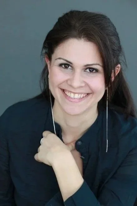 A woman with dark hair, smiling, wearing a dark shirt and long earrings, posing against a plain background.