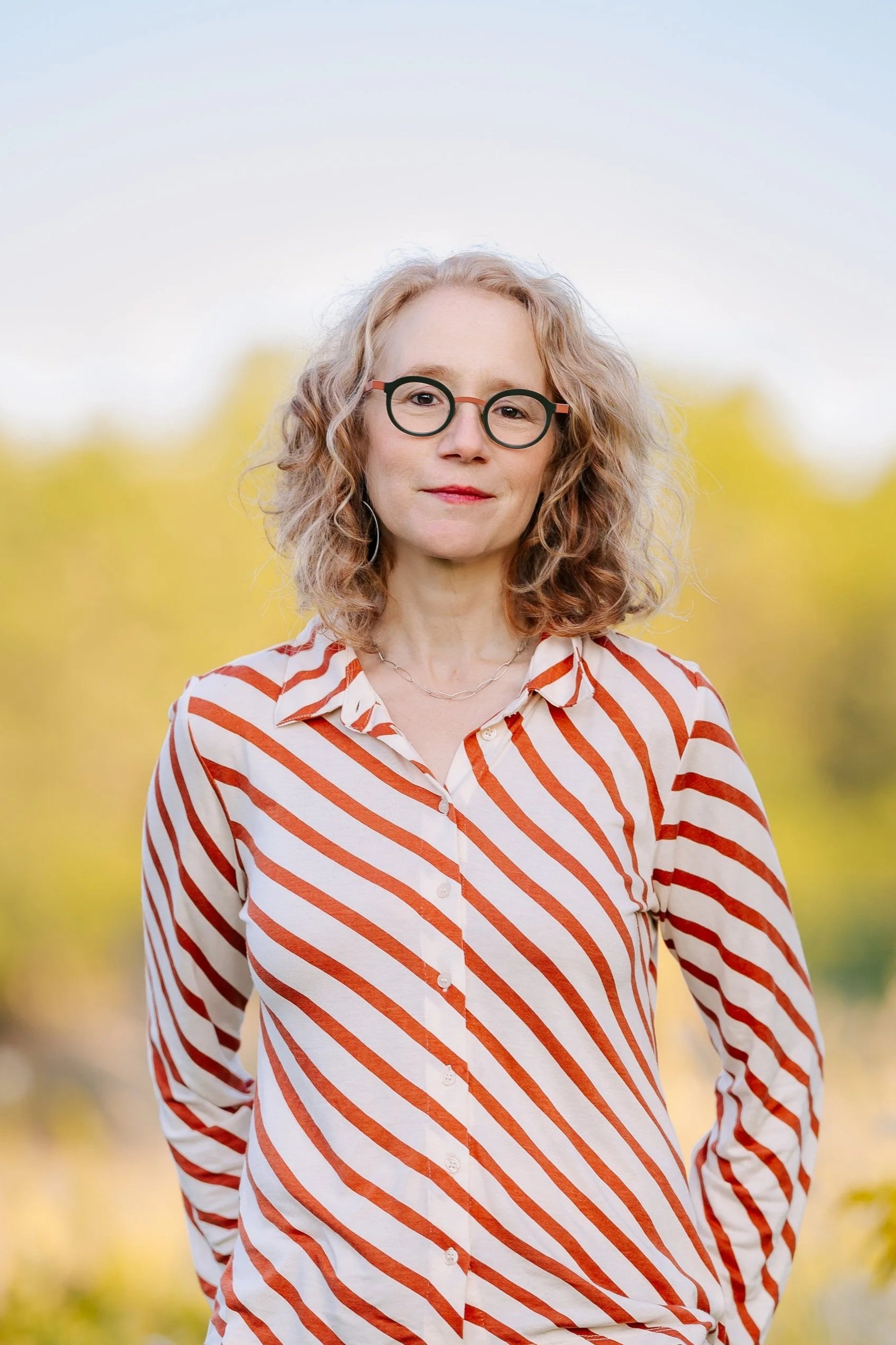 A woman with blonde curly hair, wearing glasses and a white shirt with red stripes, standing outdoors with blurred yellow and green foliage in the background.
