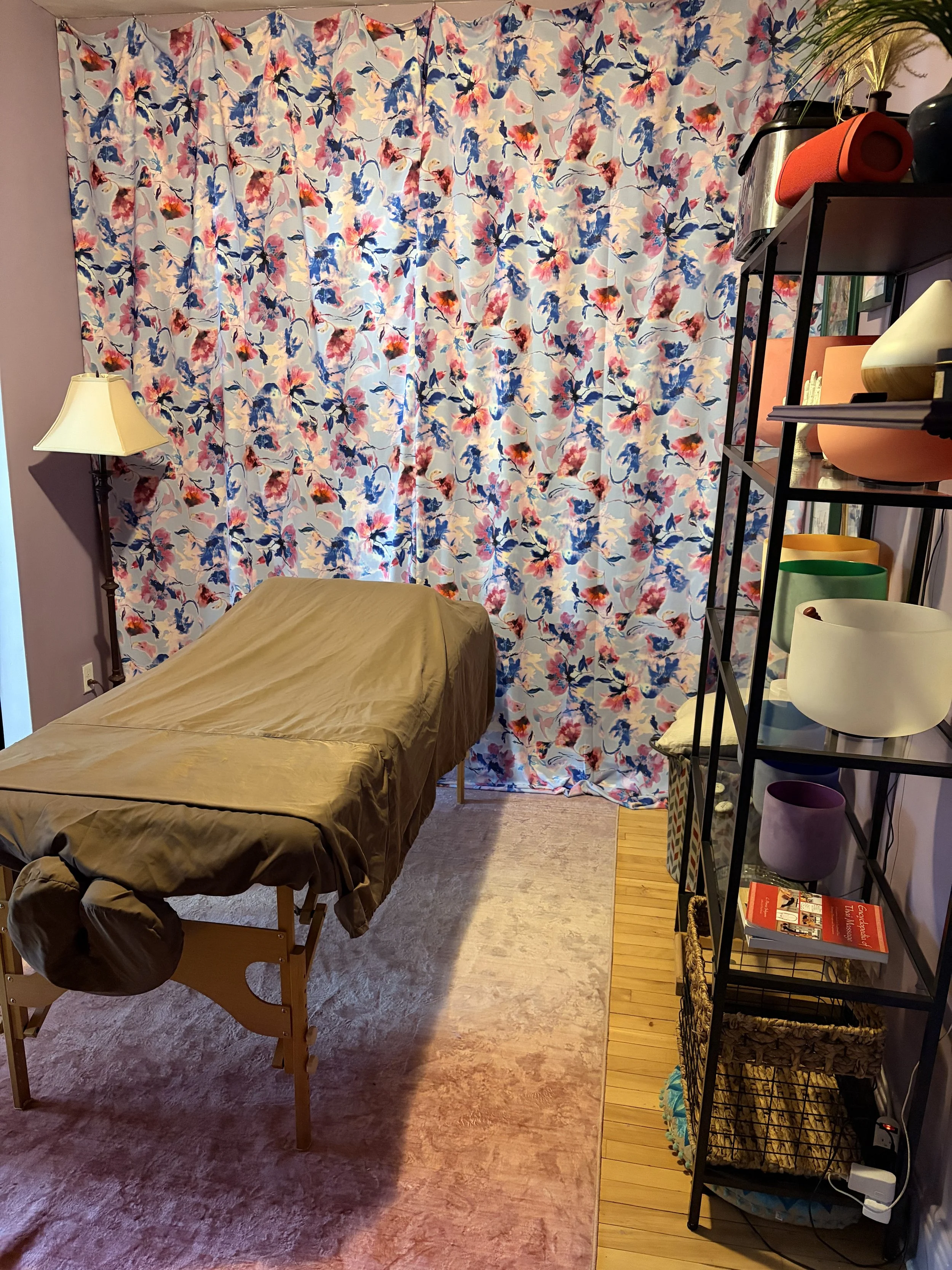 A cozy room with a massage table covered in a brown sheet, a tall lamp on the left, a floral curtain in the background, and a black metal shelf on the right holding a speaker, plants, cups, and books. The room has a soft, relaxed atmosphere suitable for therapy or relaxation.