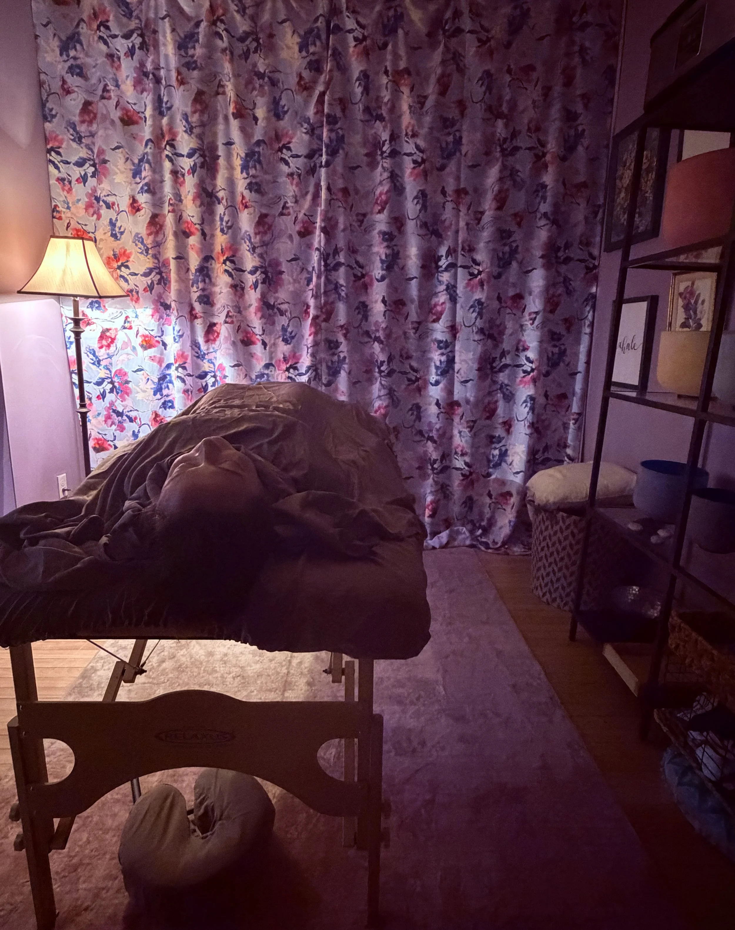 A dimly lit room with a table covered with a brown cloth, a floor lamp with a beige shade next to a wall with floral curtains, a shelf with framed pictures and books, and a brown ottoman.