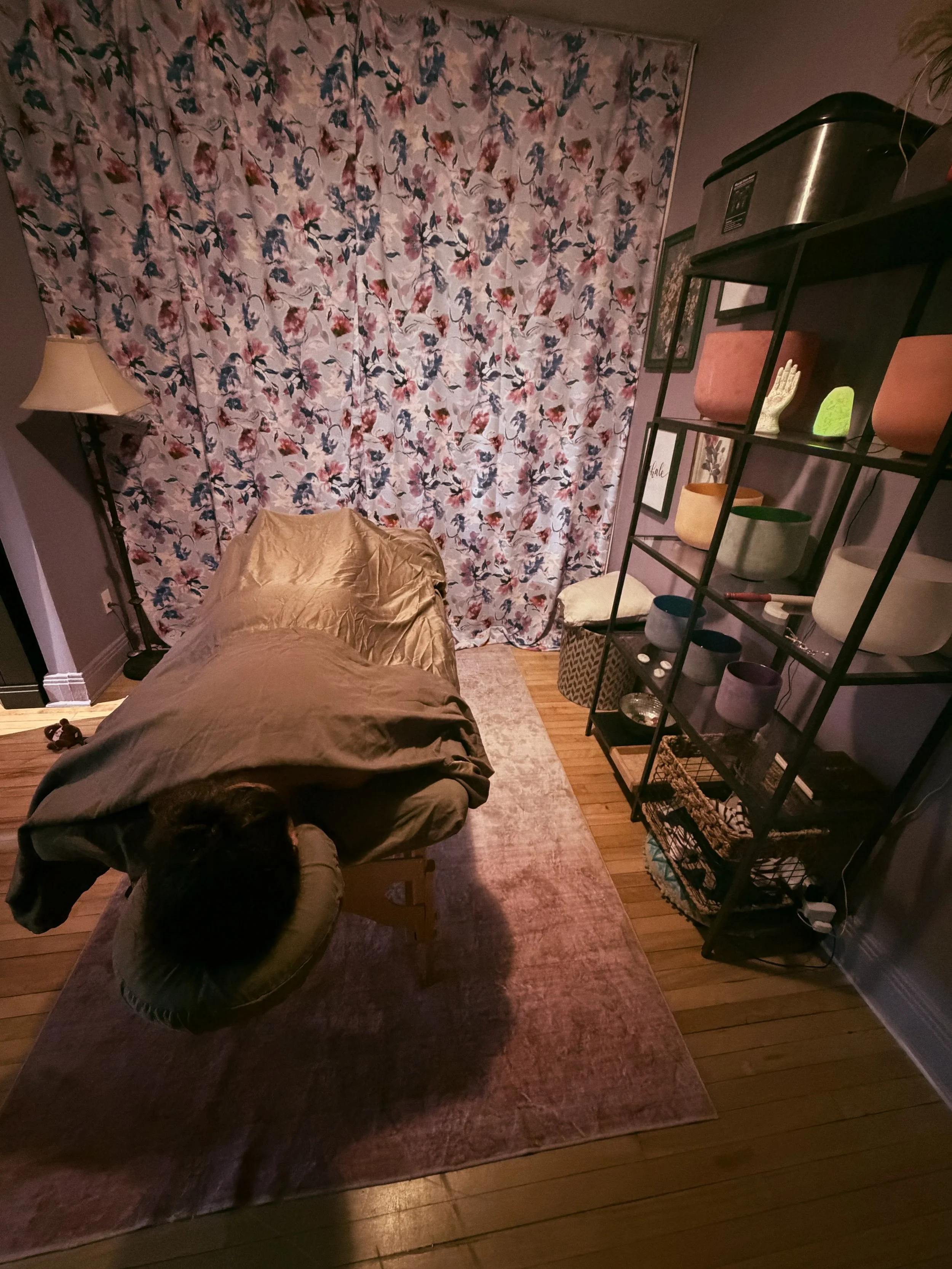 A cozy corner of a room with a floral curtain covering a window, a pinkish rug on a wooden floor, a beige chair with a blanket, a black metal shelf holding various decorative items including potted plants and framed pictures, and a small lamp on a side table. There is a person with dark hair sitting on the chair.