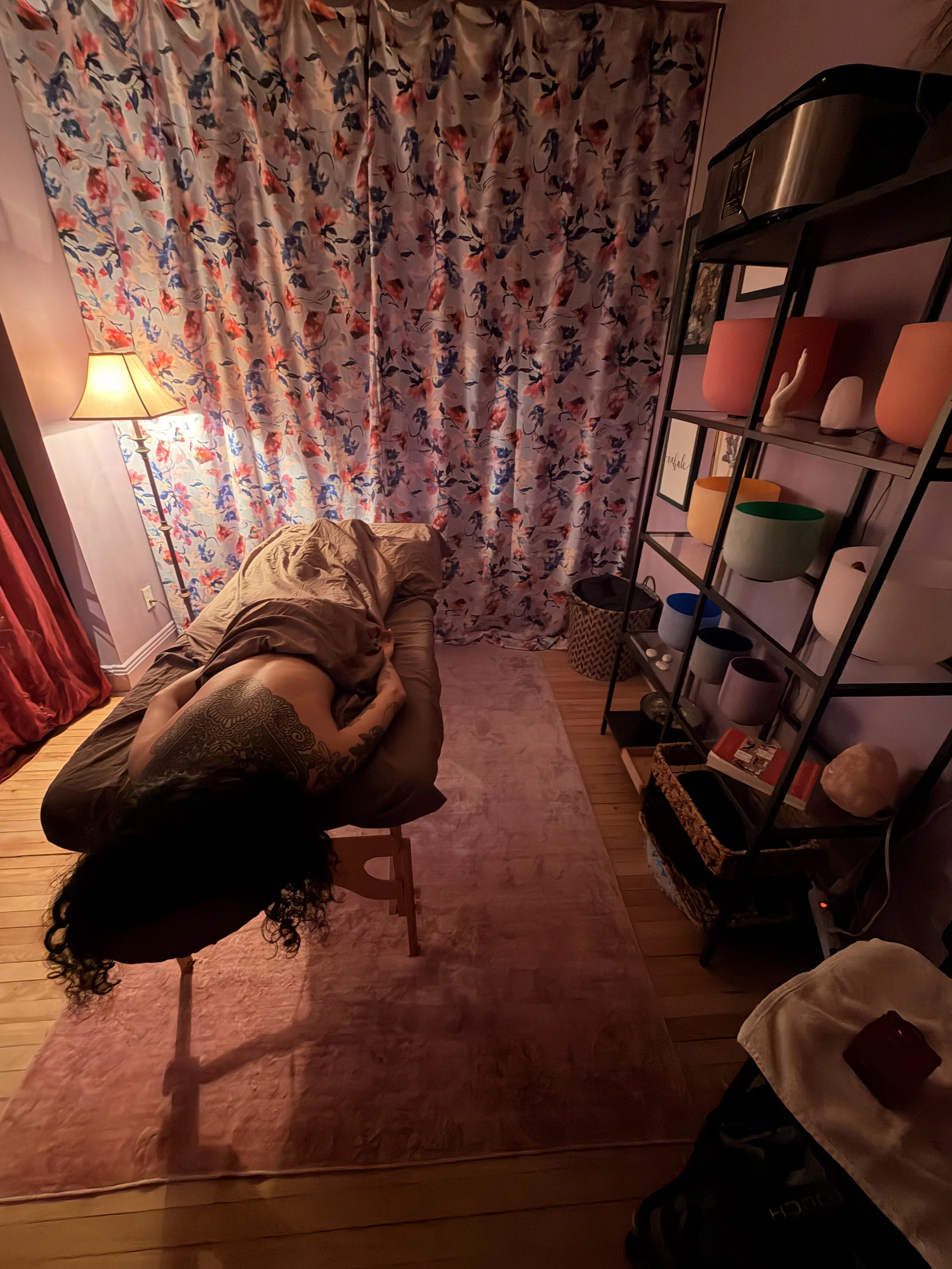 A person with curly hair lying on a bed with a brown blanket in a warmly lit room. Behind them, there is a floral curtain with red, blue, and pink patterns covering a window. To the right, a black metal shelf holds various colorful vases and decorative items, with framed pictures on the wall behind it. A small pink rug is on the wooden floor, and a floor lamp with a beige shade is turned on, illuminating the room.