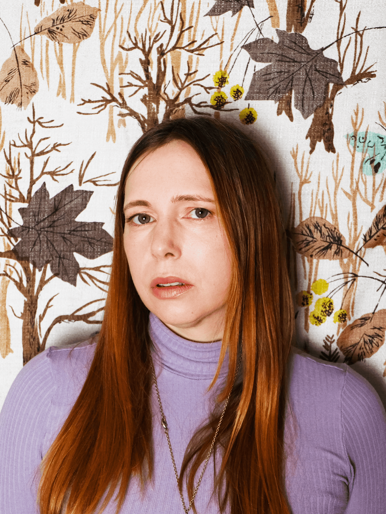A woman with long reddish-brown hair and blue eyes wearing a lavender turtleneck and a silver necklace, standing in front of a wall with an autumnal tree and leaf pattern.