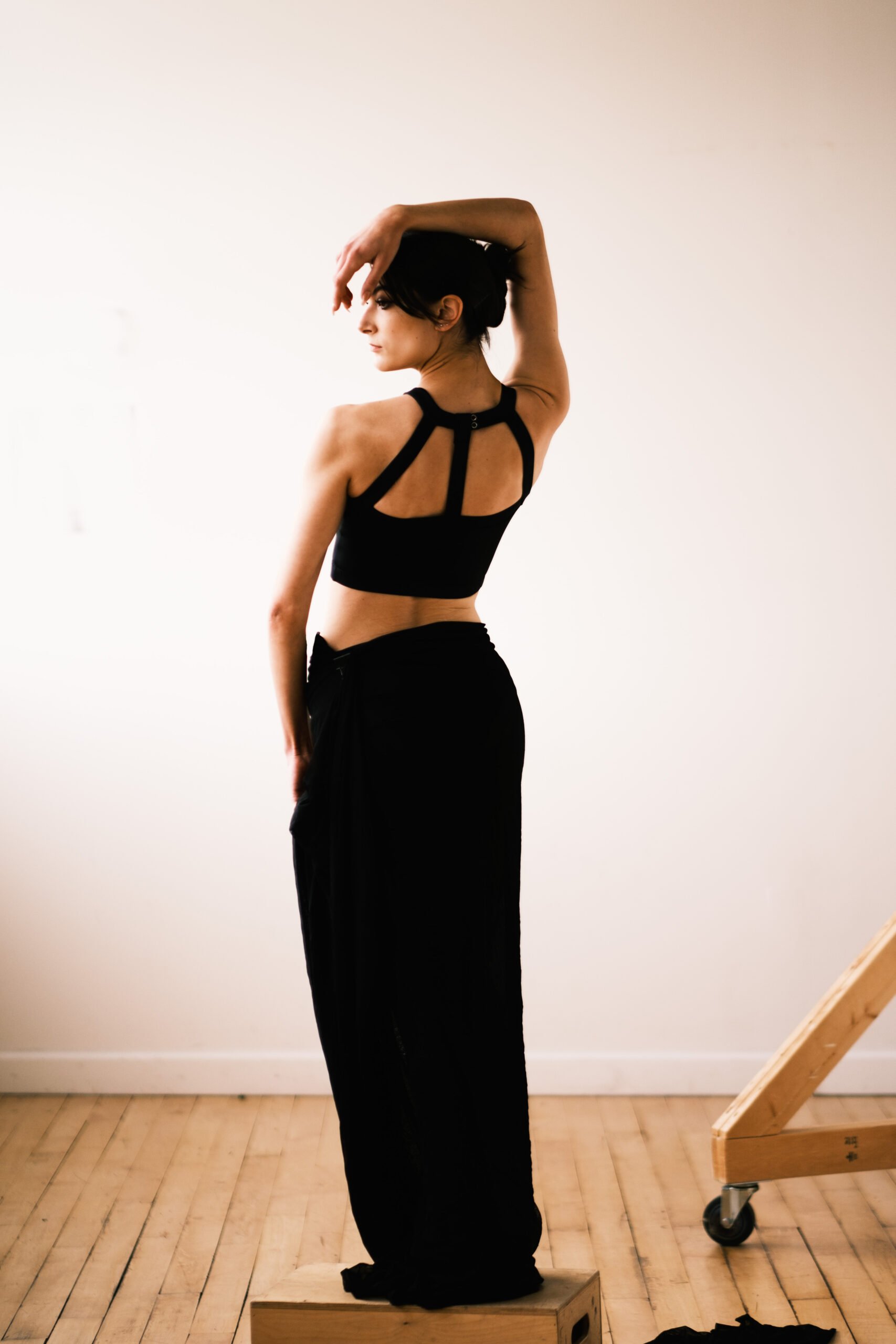 A woman with short dark hair and light skin, wearing a black sports bra and black pants, is standing with her left arm raised, resting her hand on her head in a dance or pose position. She is looking to the left with a serious expression. She is stan