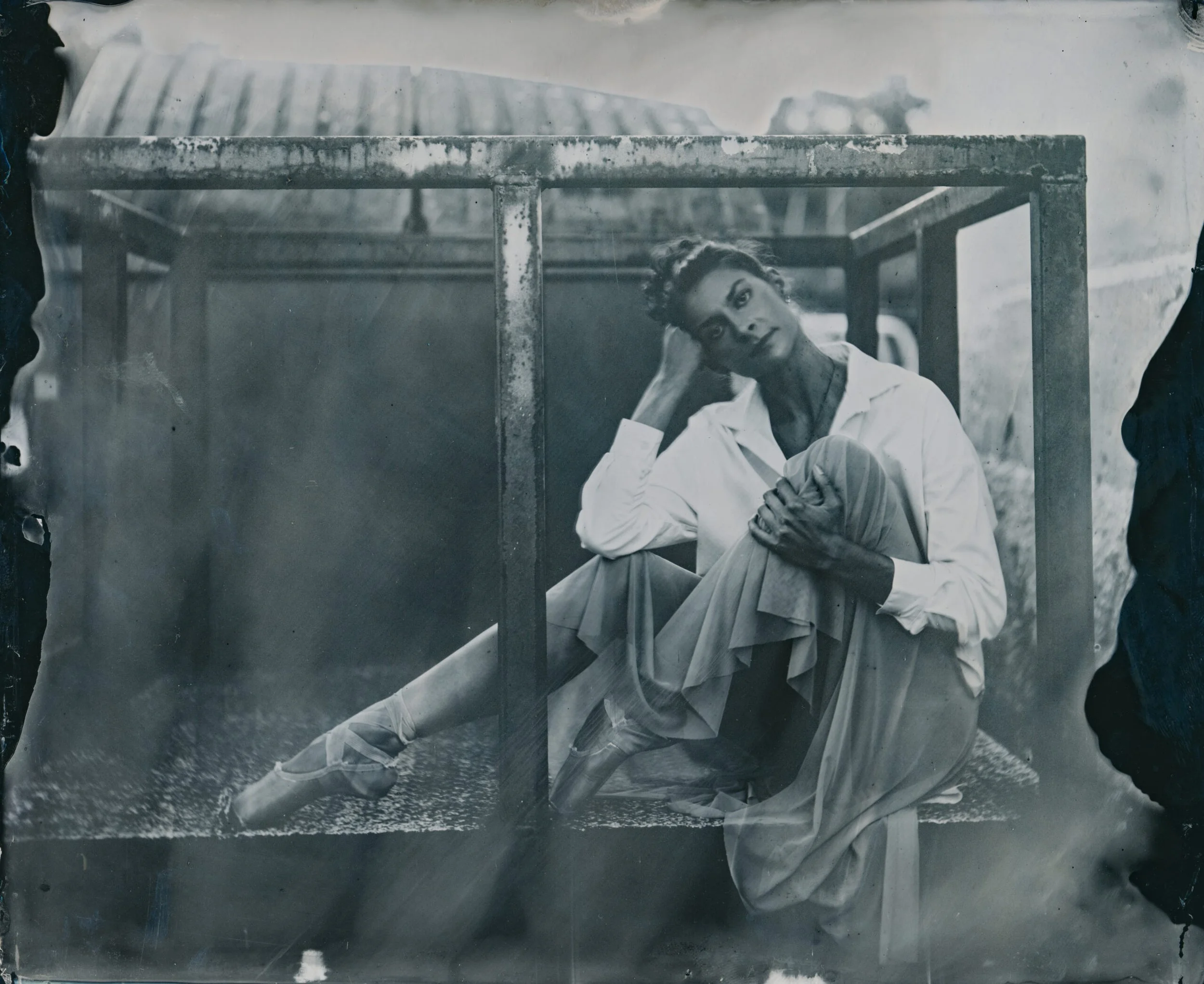 Black and white photograph of a woman with styled hair, sitting under a weathered metal structure, wearing a white blouse, looking directly at the camera.
