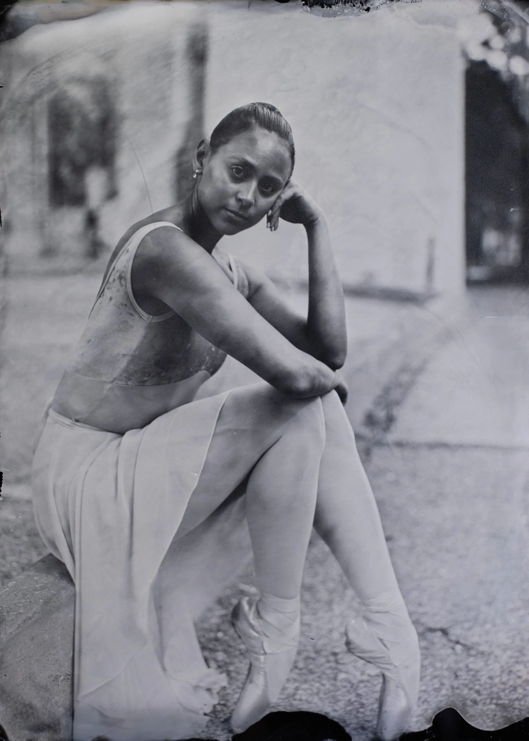 A black-and-white photograph of a woman sitting outdoors on a cloudy day. She has her hair tied back and is wearing a sleeveless top and loose pants. The woman has her left hand resting on her face and is looking at the camera.