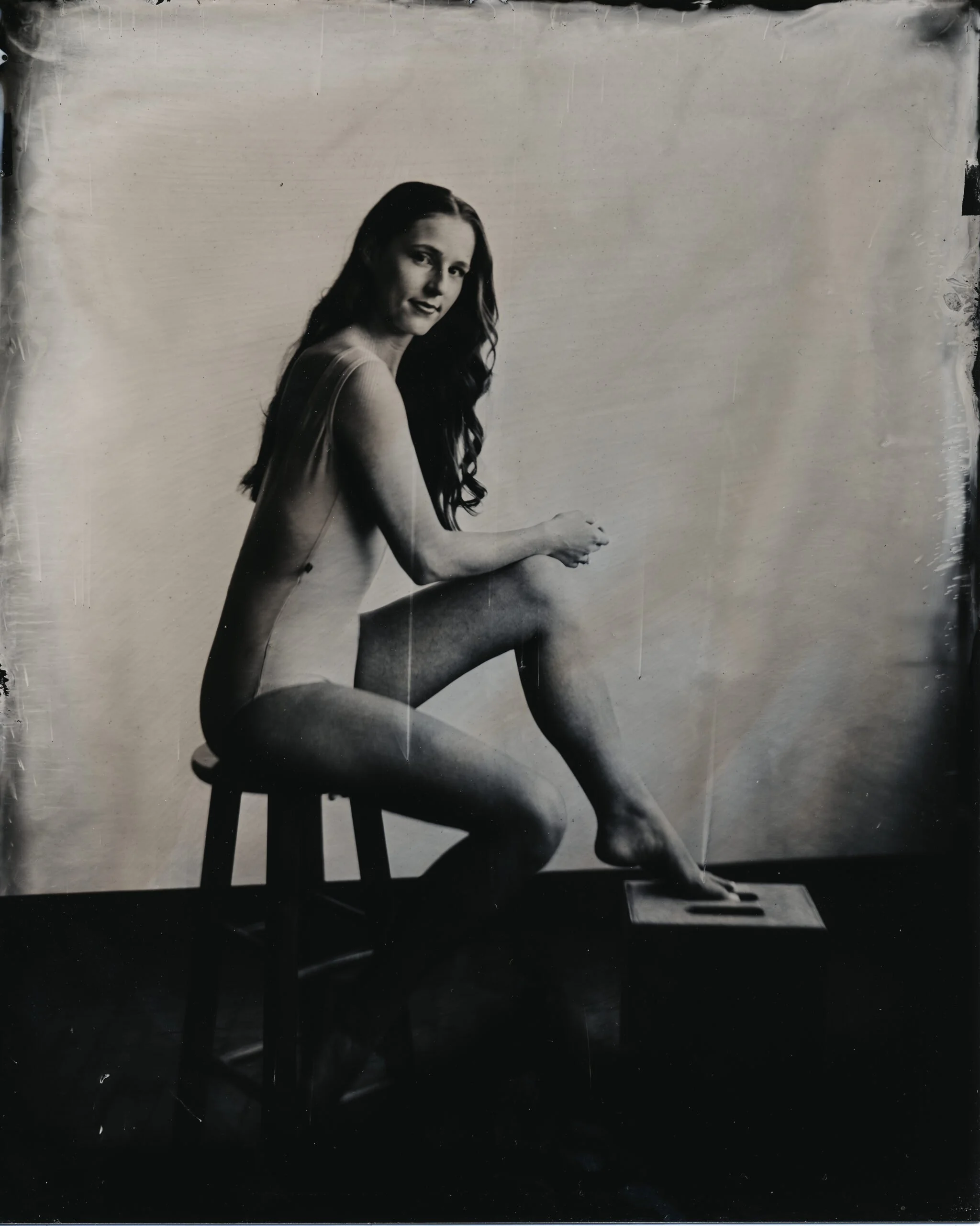 Black and white photo of a woman with long hair sitting on a chair, looking at the camera, with one knee raised and her hands resting on her knee, in front of a plain background.